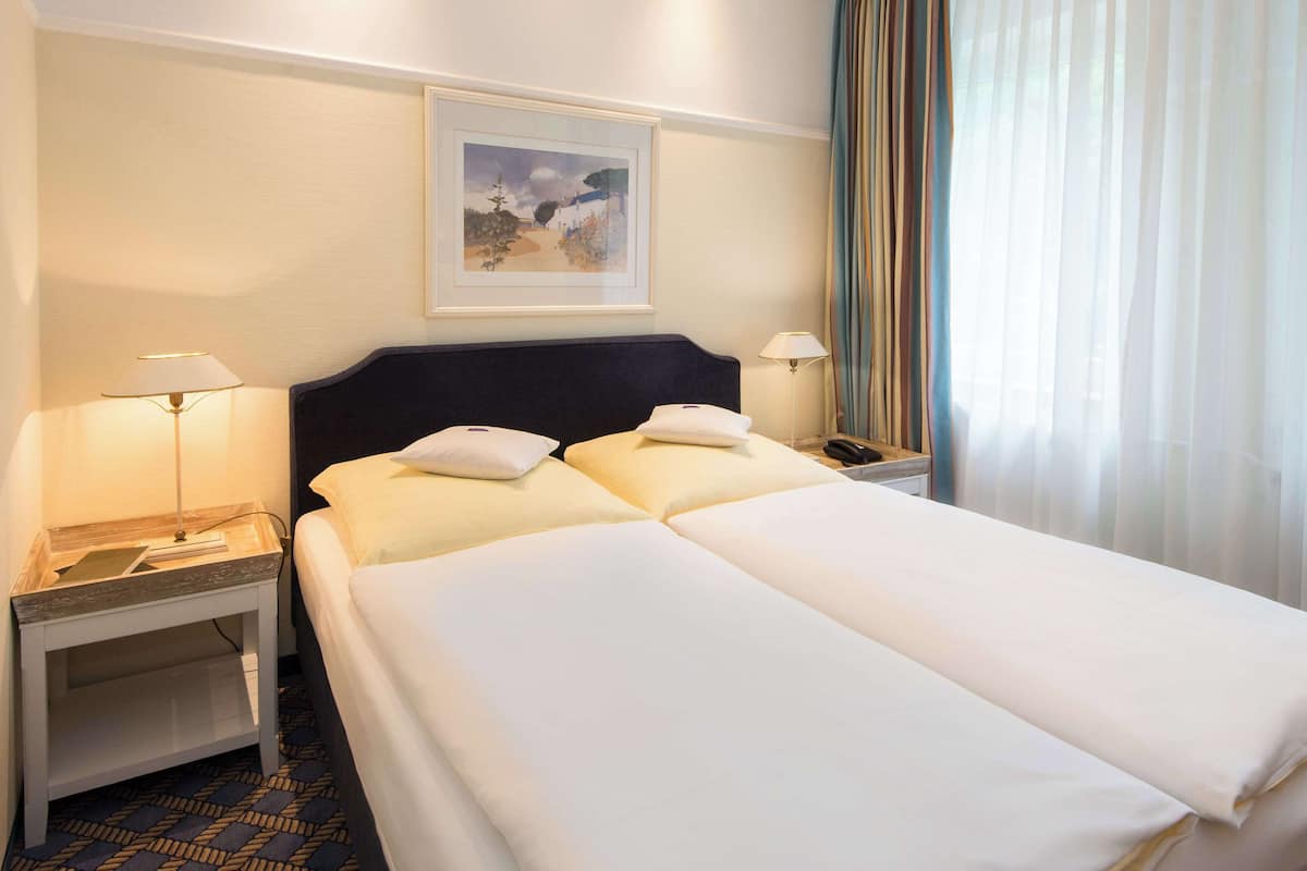 A hotel room with a double bed, two bedside tables, a nightstand, a framed picture on the wall, and a window with curtains.