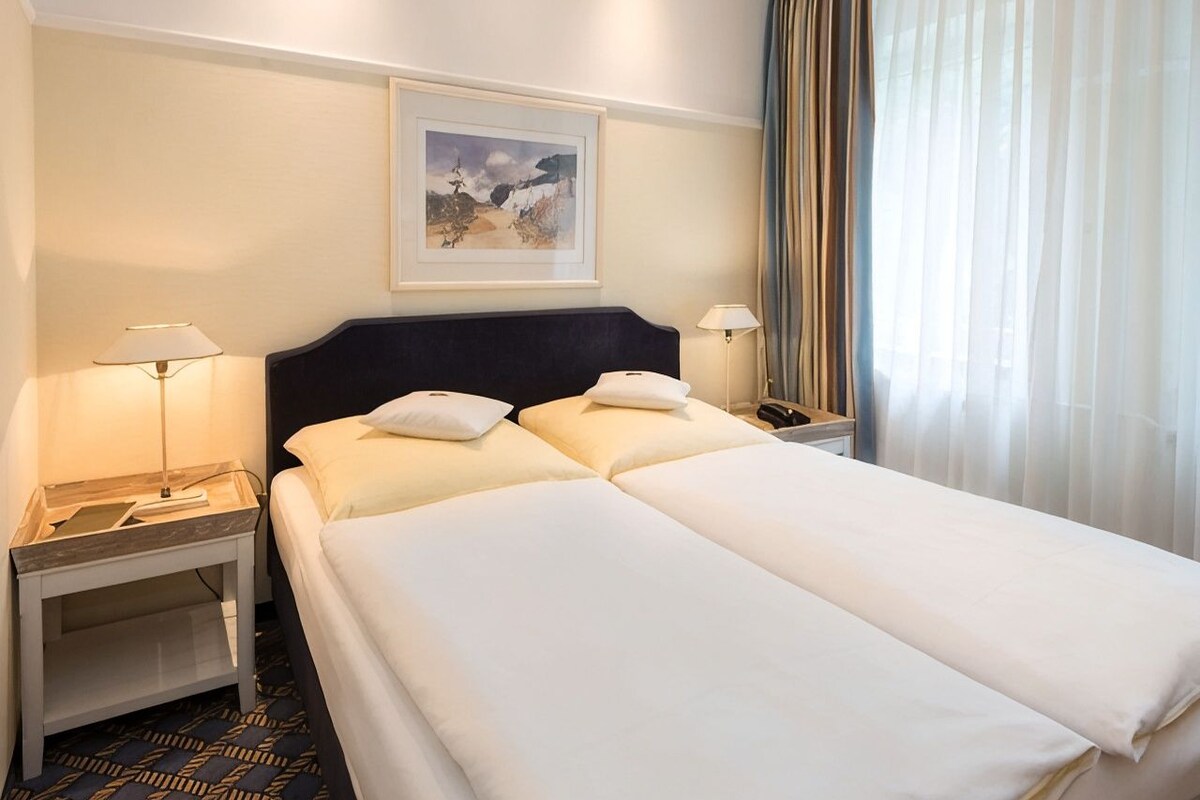 Quarto de hotel com cama de casal, duas mesas de cabeceira com abajures, uma obra de arte emoldurada na parede e uma janela com cortinas.