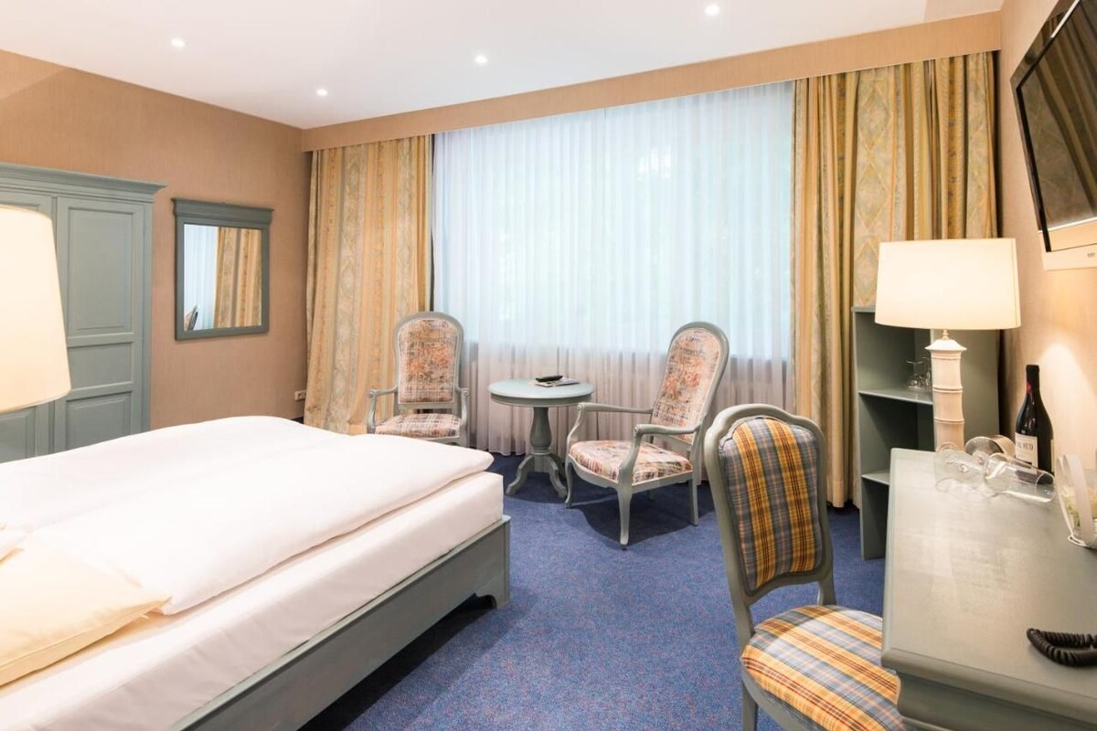 Quarto de hotel com cama, duas cadeiras, mesa redonda pequena, escrivaninha com luminária e televisão.