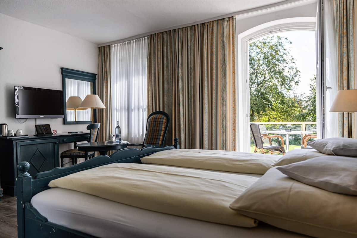 A hotel room with two beds, a desk with a TV, lamps, and a view of greenery through the window.