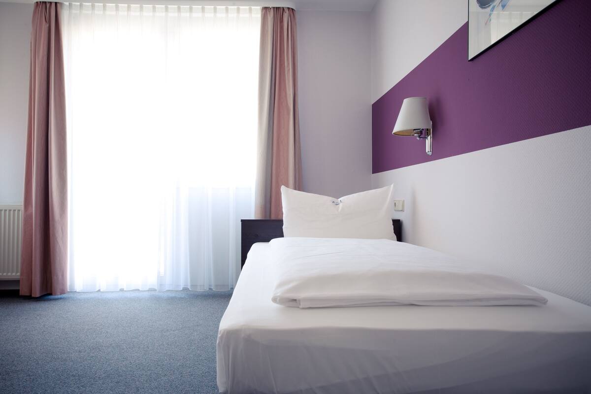 A hotel room with a bed, a nightstand, a wall-mounted lamp, and a window with curtains.