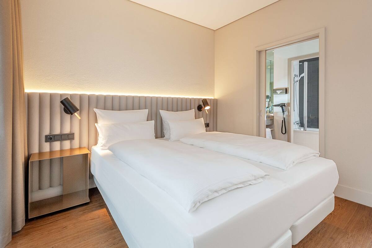 A modern hotel room with a large bed, a bedside table, a nightstand, and a wall-mounted lamp.