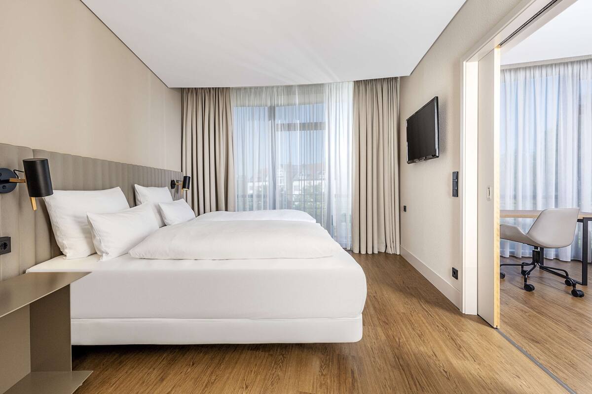 A modern hotel room with a large bed, a desk, a chair, and a TV on the wall.