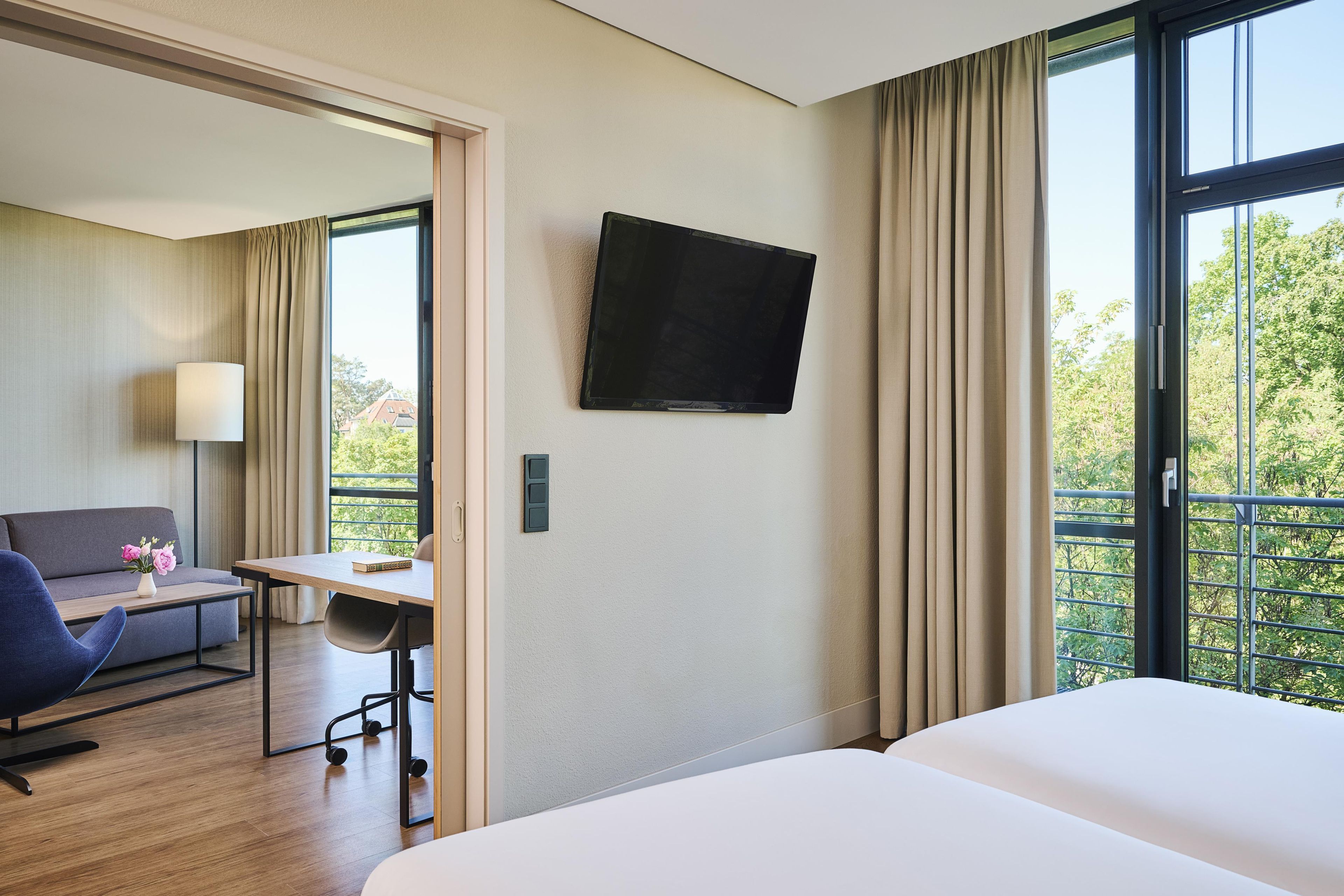 Ein Hotelzimmer mit einem Bett, einem Schreibtisch mit Stuhl, einer Couch, einem an der Wand befestigten Fernseher und einem Fenster mit Vorhängen.