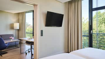 Ein Hotelzimmer mit einem Bett, einem Schreibtisch mit Stuhl, einer Couch, einem an der Wand befestigten Fernseher und einem Fenster mit Vorhängen.