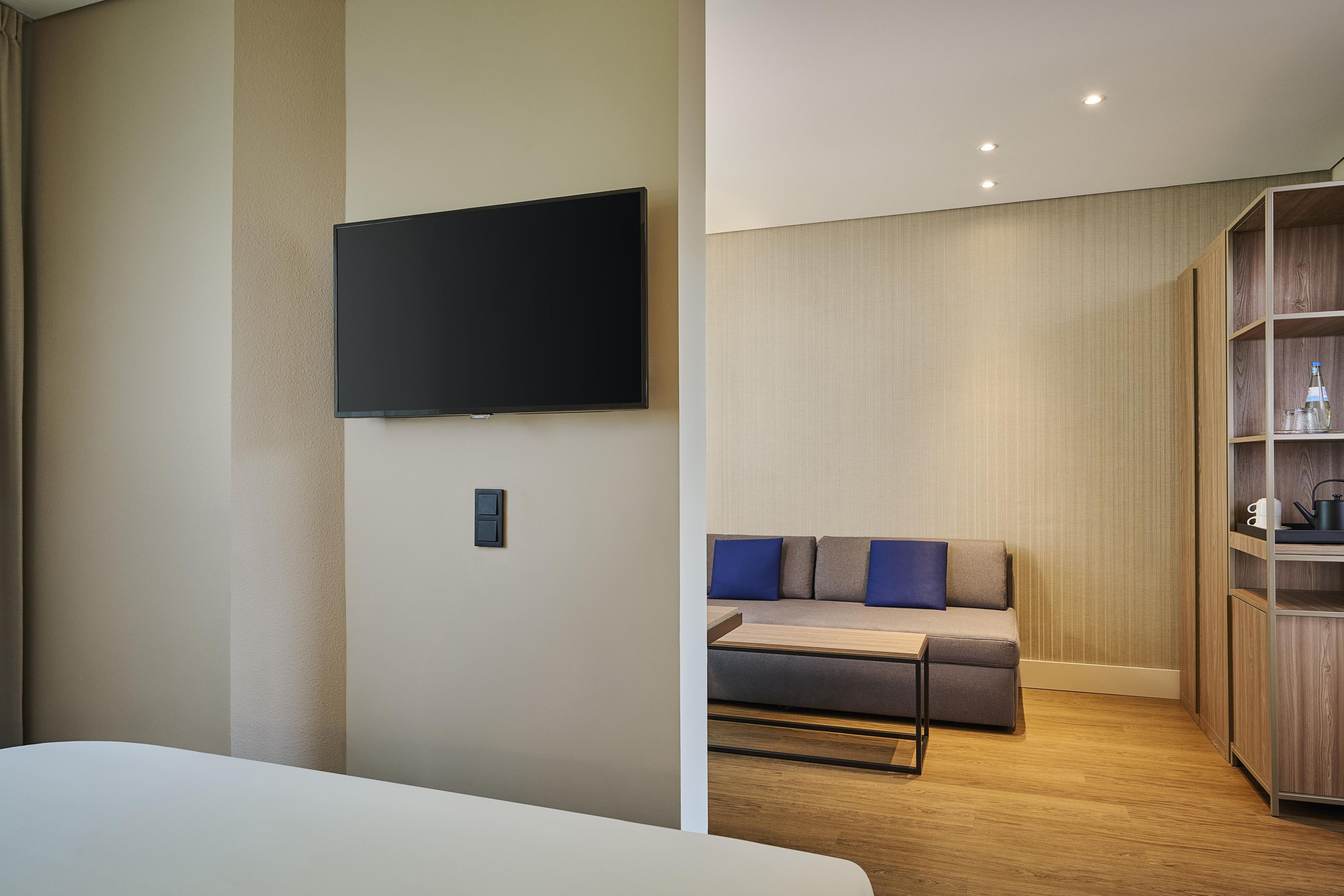 Una habitación de hotel moderna con un televisor de pantalla plana en la pared, un sofá con cojines azules, piso de madera y un mueble de madera empotrado.