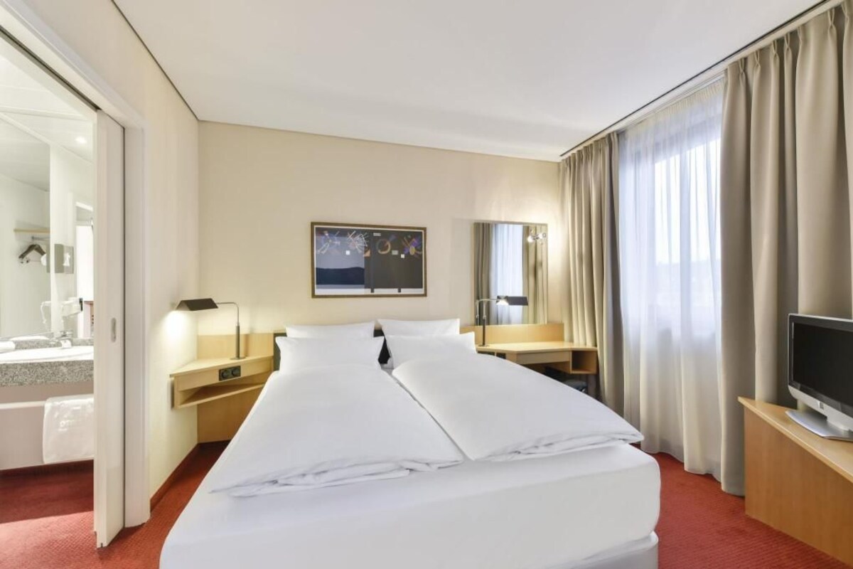 Habitación de hotel con cama, escritorio, televisión, espejo y ventana con cortinas.