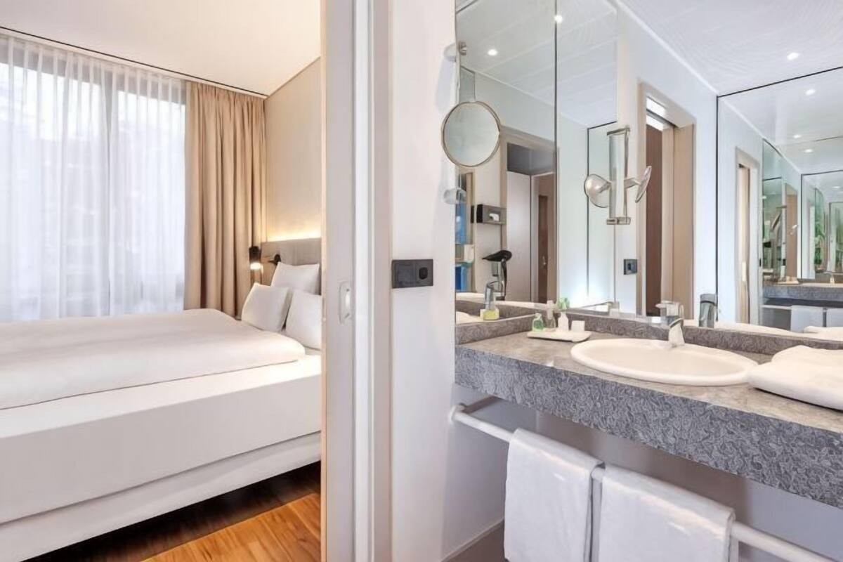 A hotel room with a bed, a large mirror, a bathroom with a sink and mirror, and a view of the outdoors.