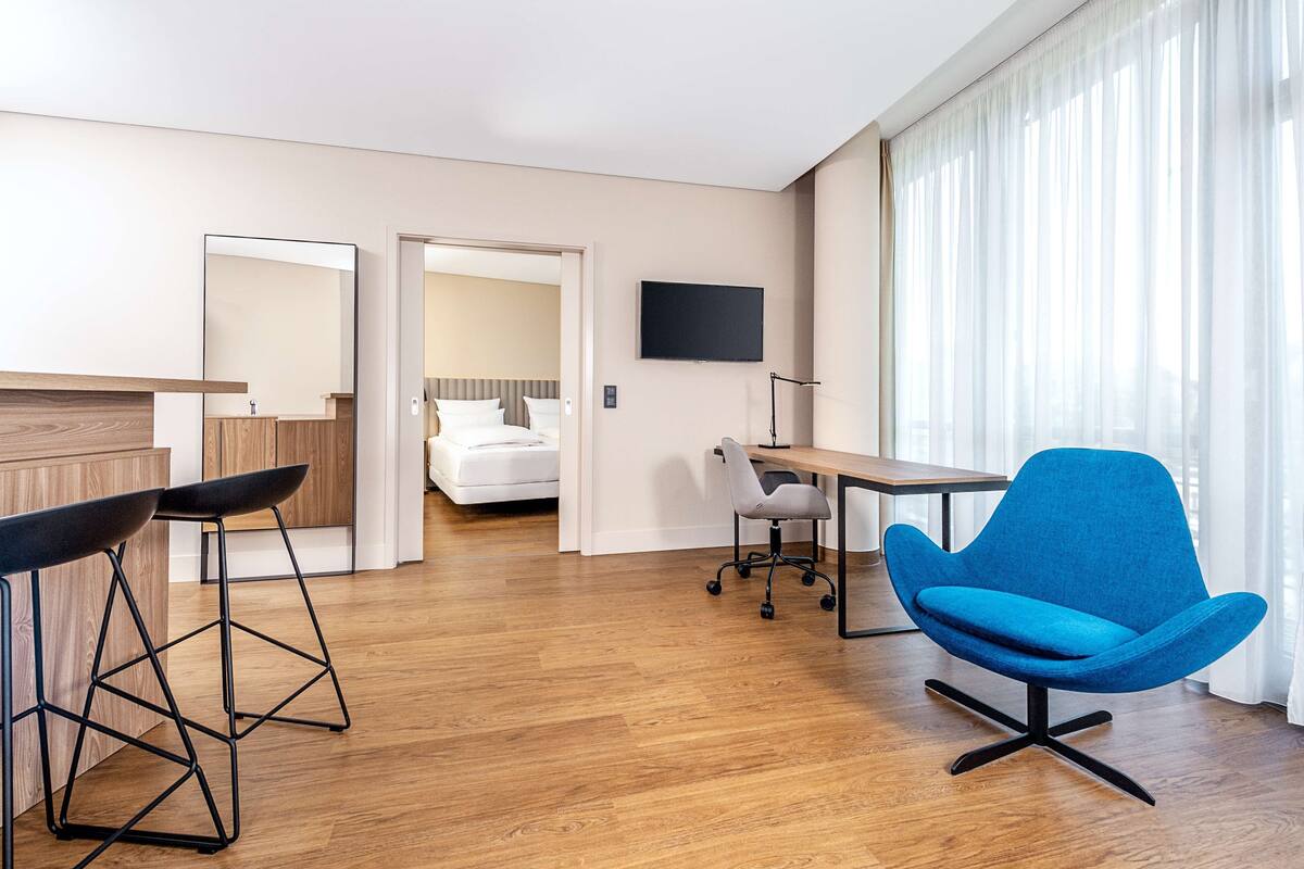 A modern hotel room with a wooden floor, a desk, a blue chair, a TV, and a bed in the background.