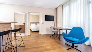 A modern hotel room with a wooden floor, a desk, a blue chair, a TV, and a bed in the background.
