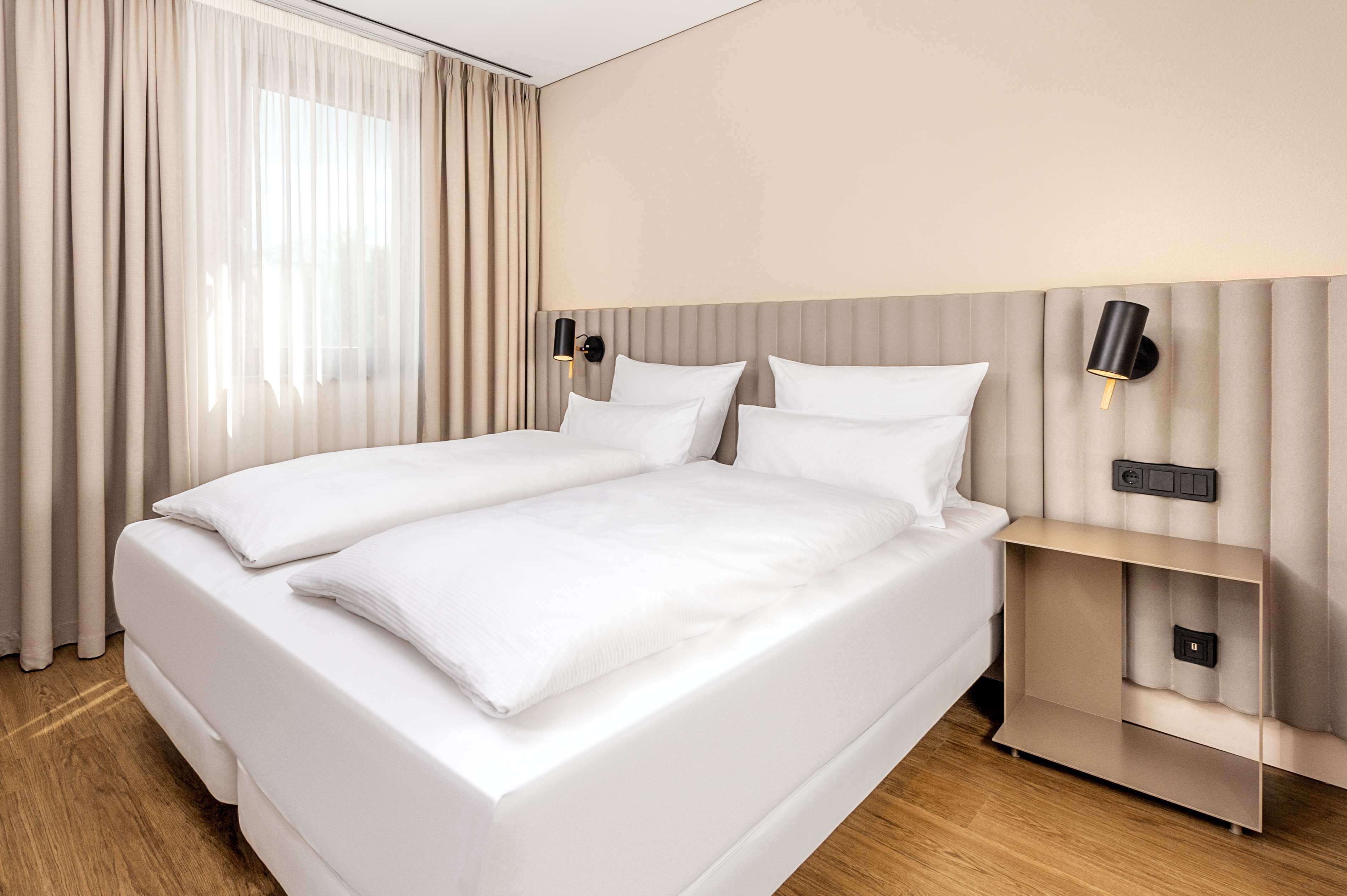 A neatly made double bed with white linens, a bedside table with a lamp, and a wall-mounted control panel.