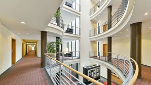 A multi-level hotel interior with a central spiral staircase, multiple balconies, and a long corridor leading to a reception area. - NH München Unterhaching (Unterhaching)
