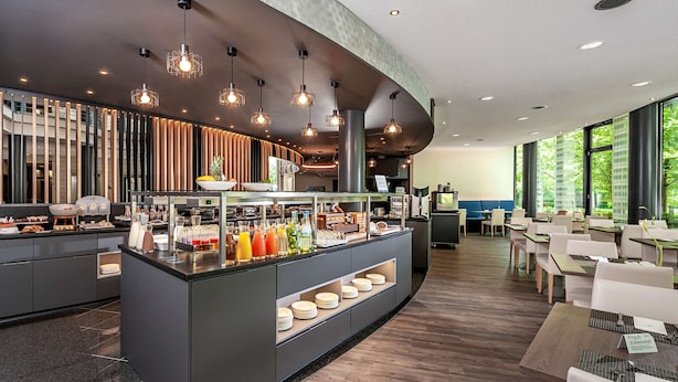 A modern hotel lobby with a breakfast bar, seating area, and large windows. - NH München Unterhaching (Unterhaching)