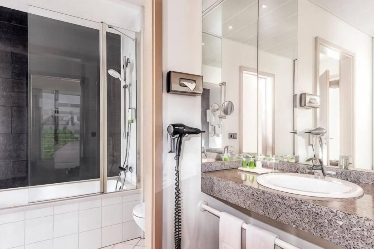 A modern bathroom with a glass shower enclosure, a built-in hairdryer, a large mirror, and a granite countertop sink.