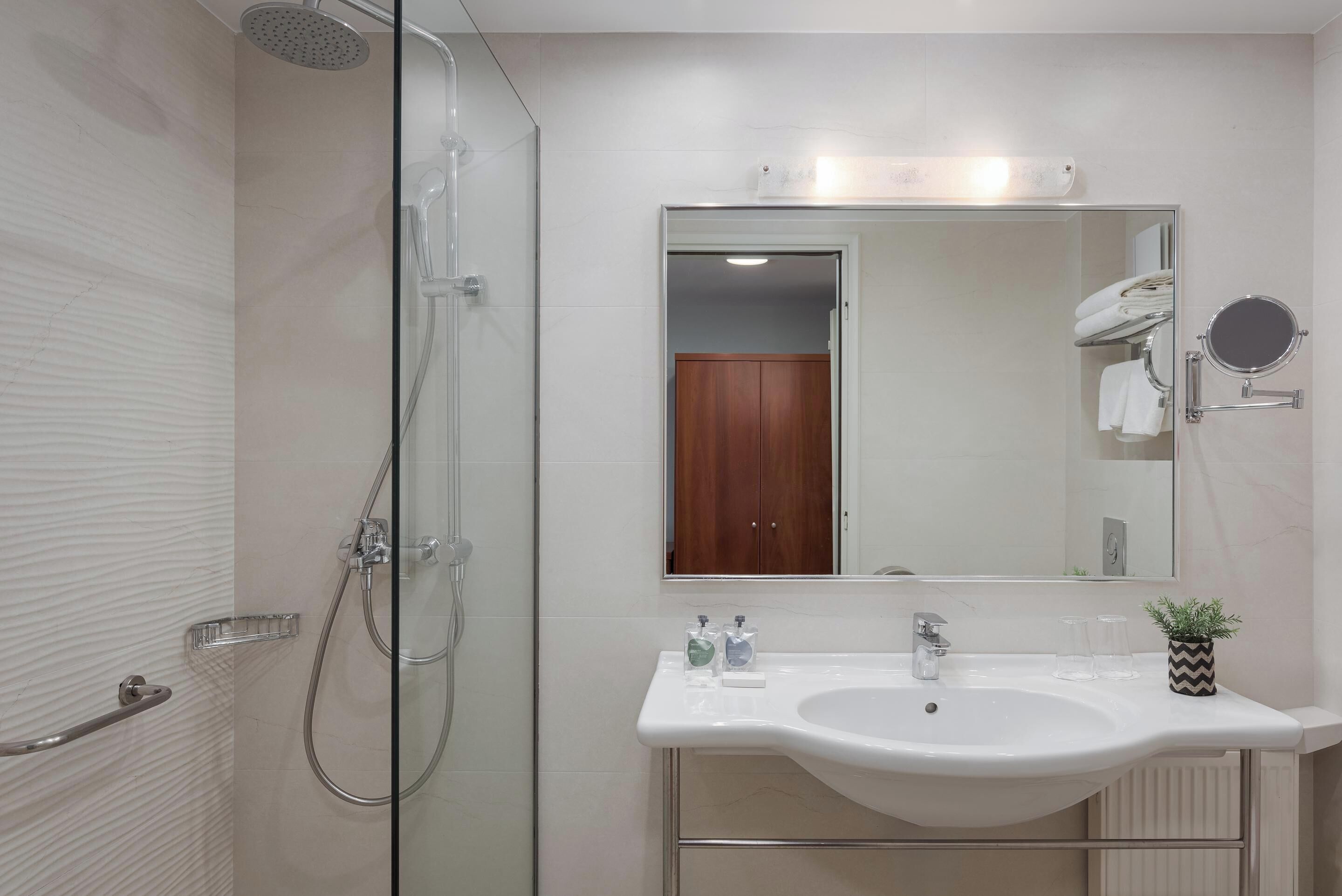 Un baño moderno con cabina de ducha de vidrio, un lavamanos blanco con un espejo encima y una pequeña planta en maceta en el borde del lavamanos.