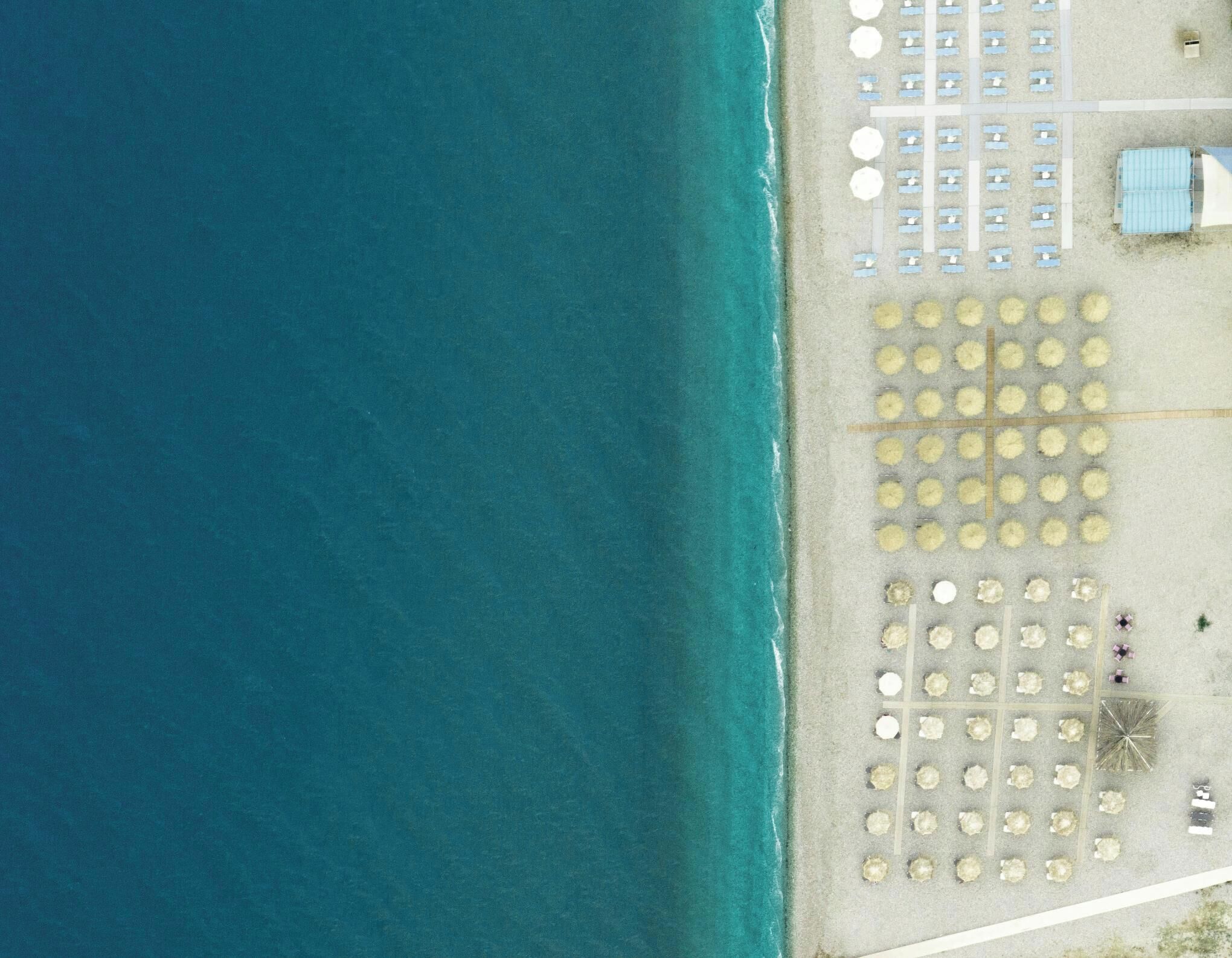 An aerial view of a beach with rows of sun loungers and umbrellas, a small building, and a clear blue sea.