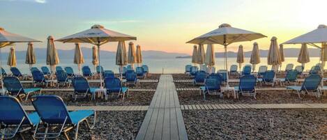 A beachside promenade with rows of blue lounge chairs and umbrellas, leading to a calm sea.