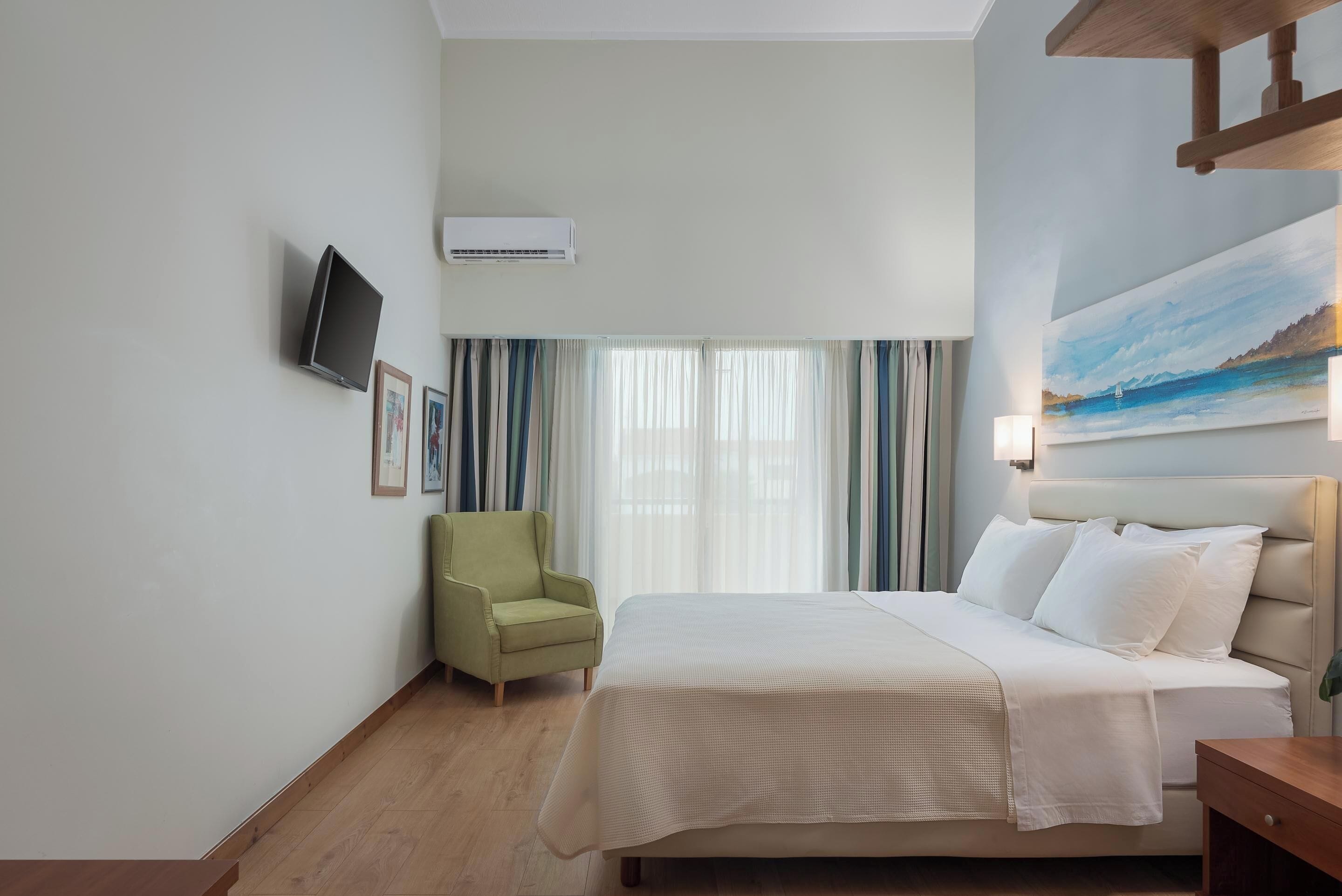 A hotel room with a bed, a green armchair, a flat-screen TV, and a painting on the wall.
