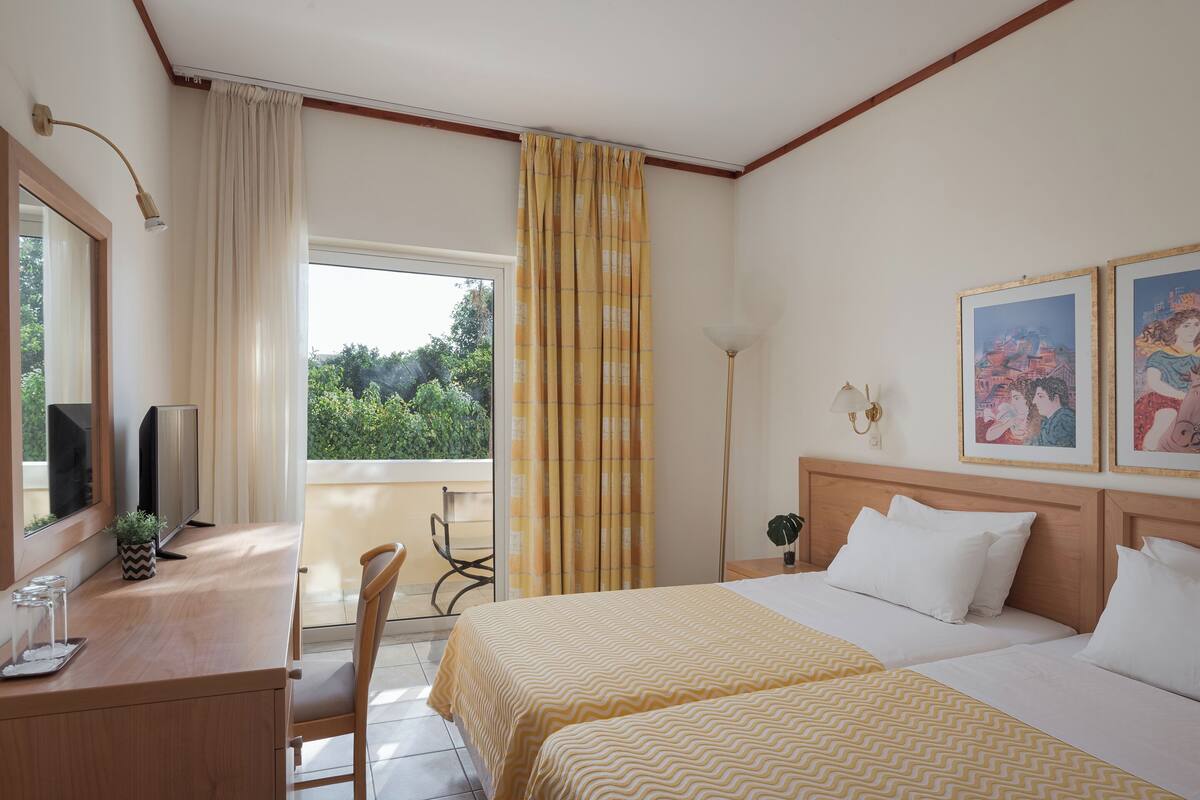 A hotel room with a bed, a desk with a TV, a chair, a window with curtains, and a balcony.