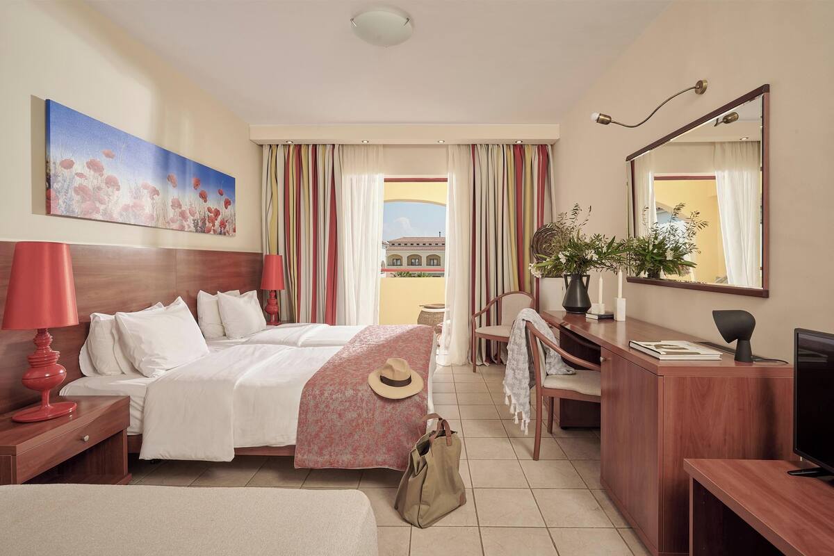 A hotel room with a bed, bedside table, lamp, desk, chair, TV, and a large window with curtains.