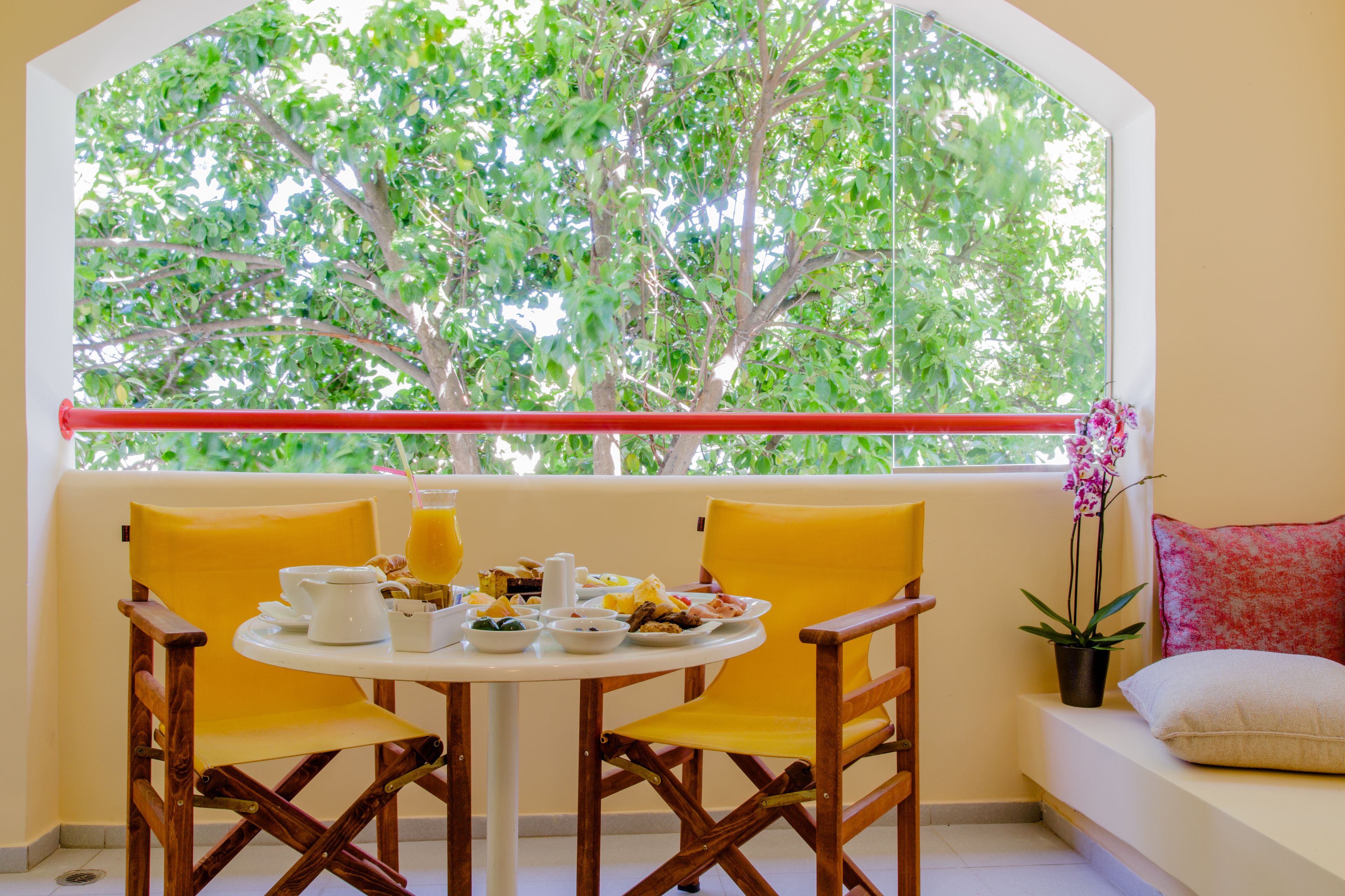 A dining area with a table set for breakfast, two yellow chairs, a window with a view of trees, and a potted plant on a bench.