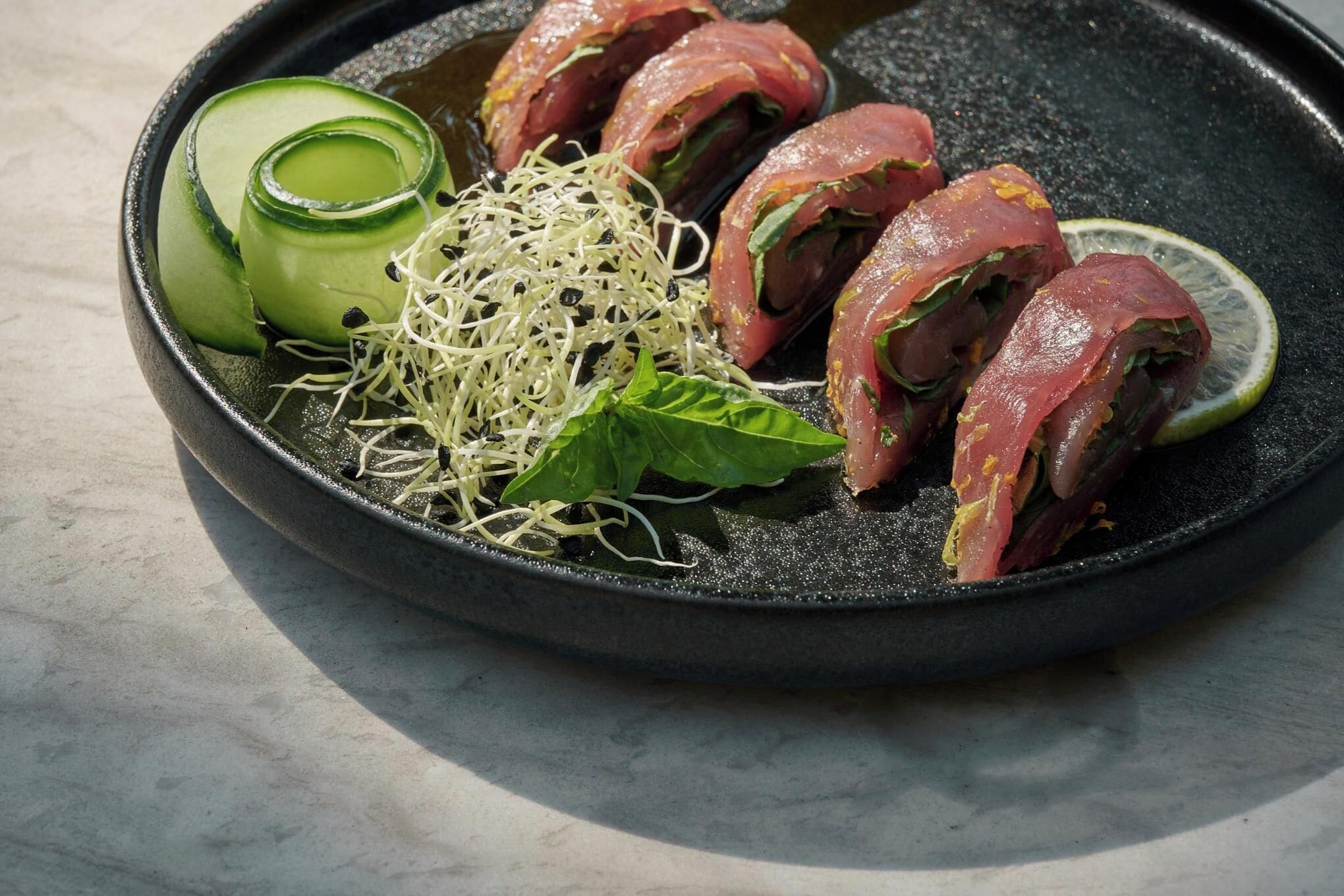 A dish with rolled meat, cucumber slices, sprouts, and a basil leaf on a black plate.