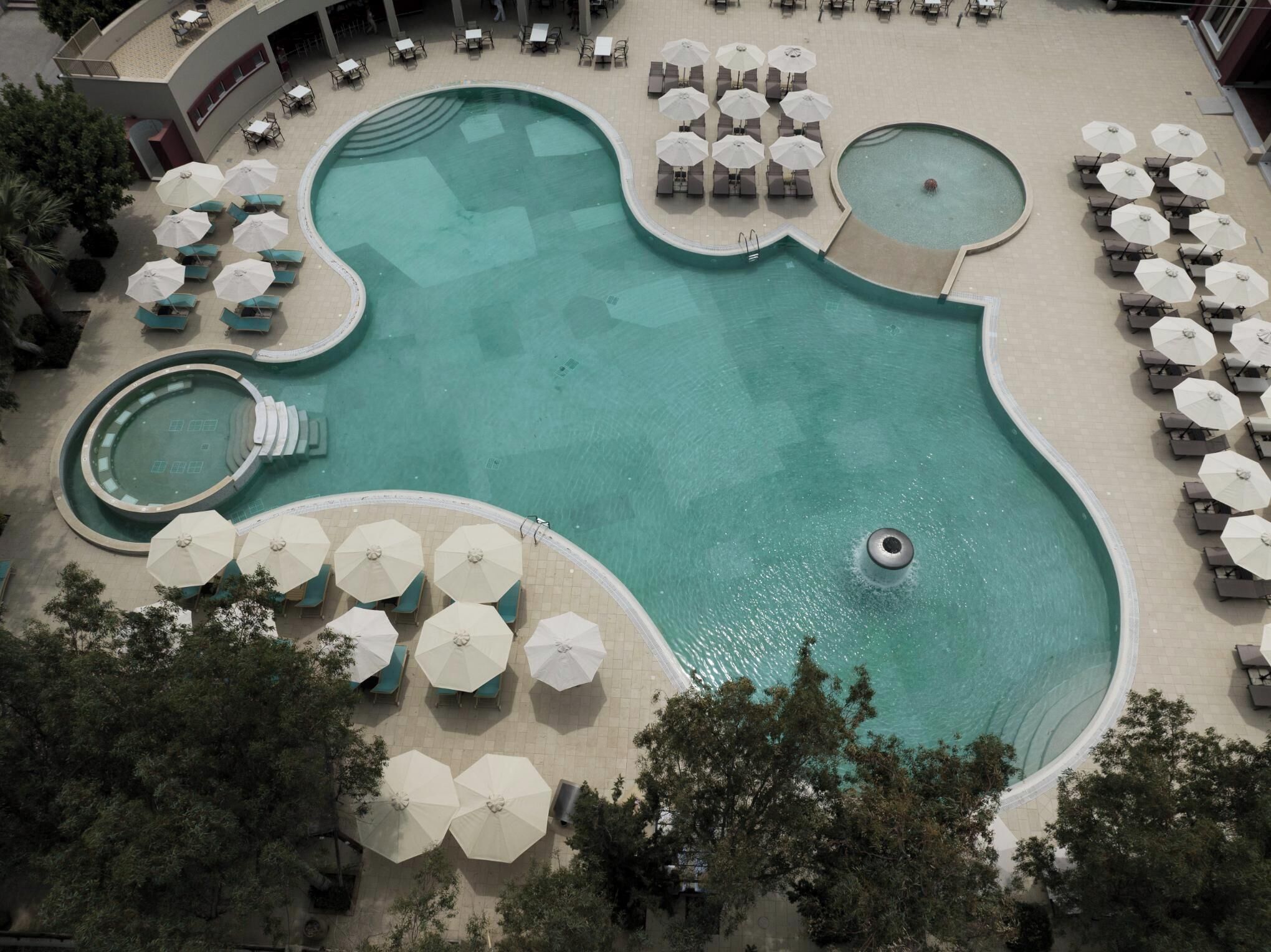 An aerial view of a pool area with multiple seating areas, including sun loungers and umbrellas, and a central swimming pool with a circular hot tub.