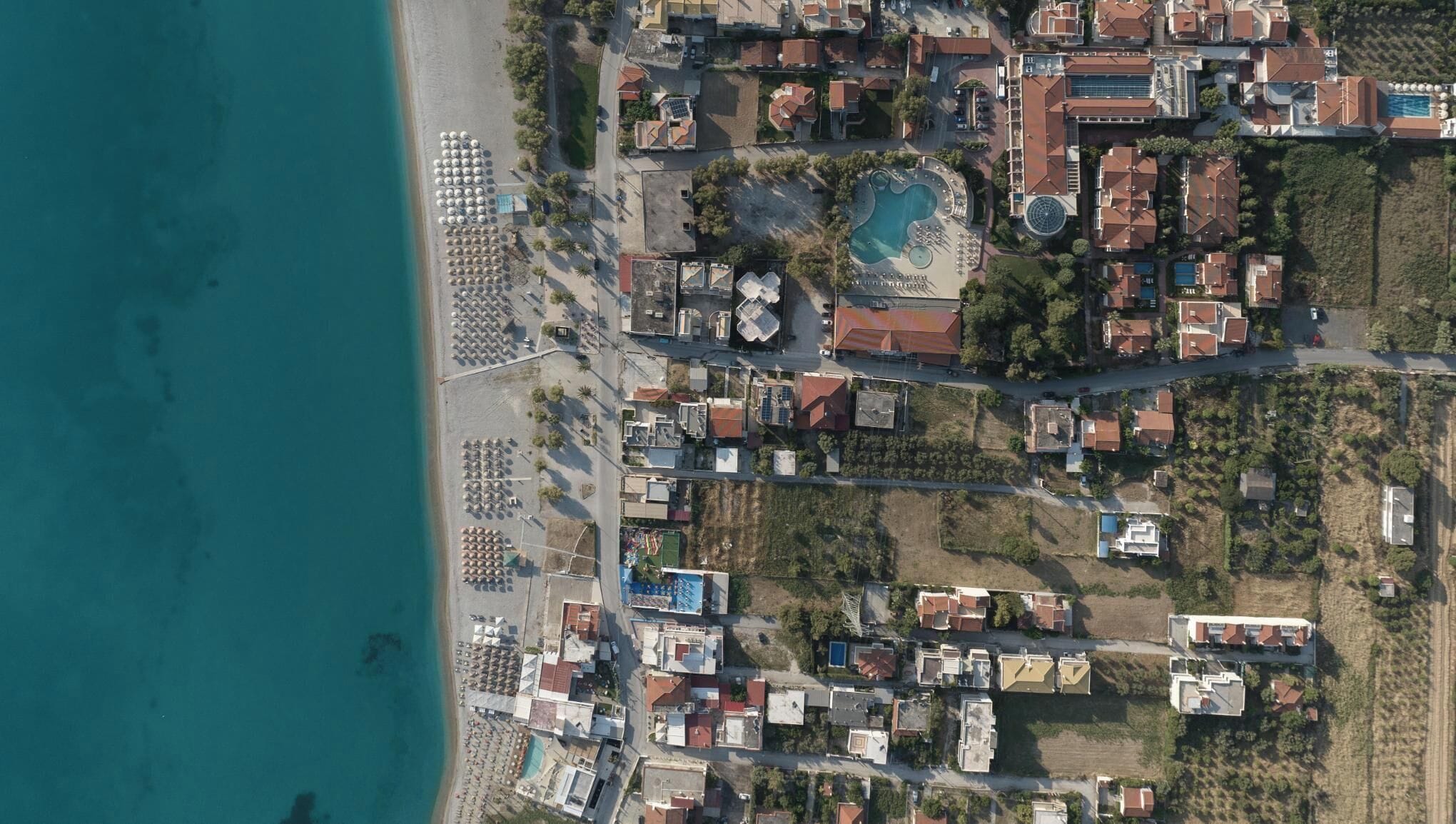 An aerial view of a coastal resort with multiple buildings, a large swimming pool, and a beach area with sun loungers.