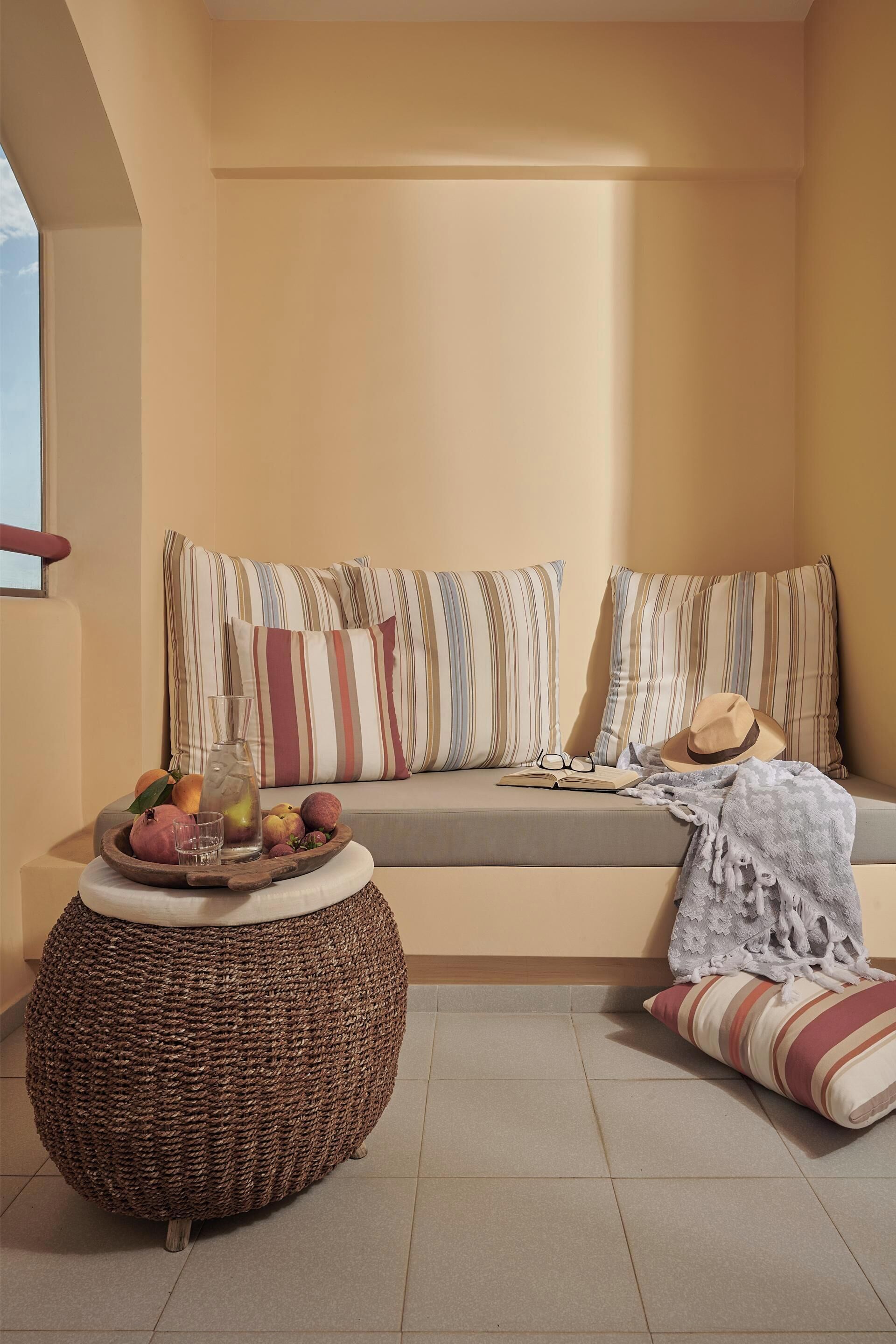 Un coin salon accueillant avec un canapé, des coussins à rayures, un pouf en osier et un plateau contenant des fruits et un livre.