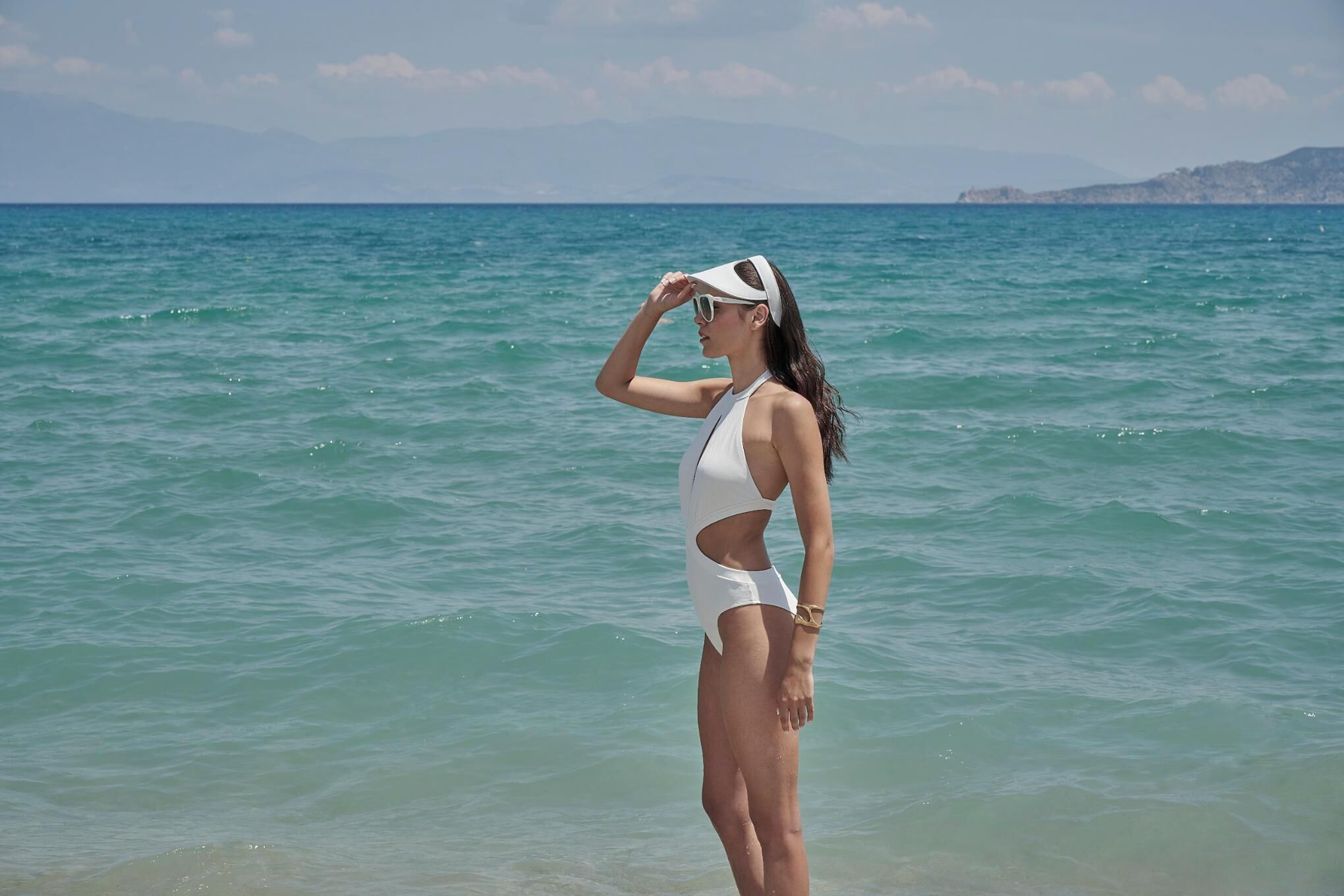 A person standing by the sea wearing a white swimsuit and a visor.