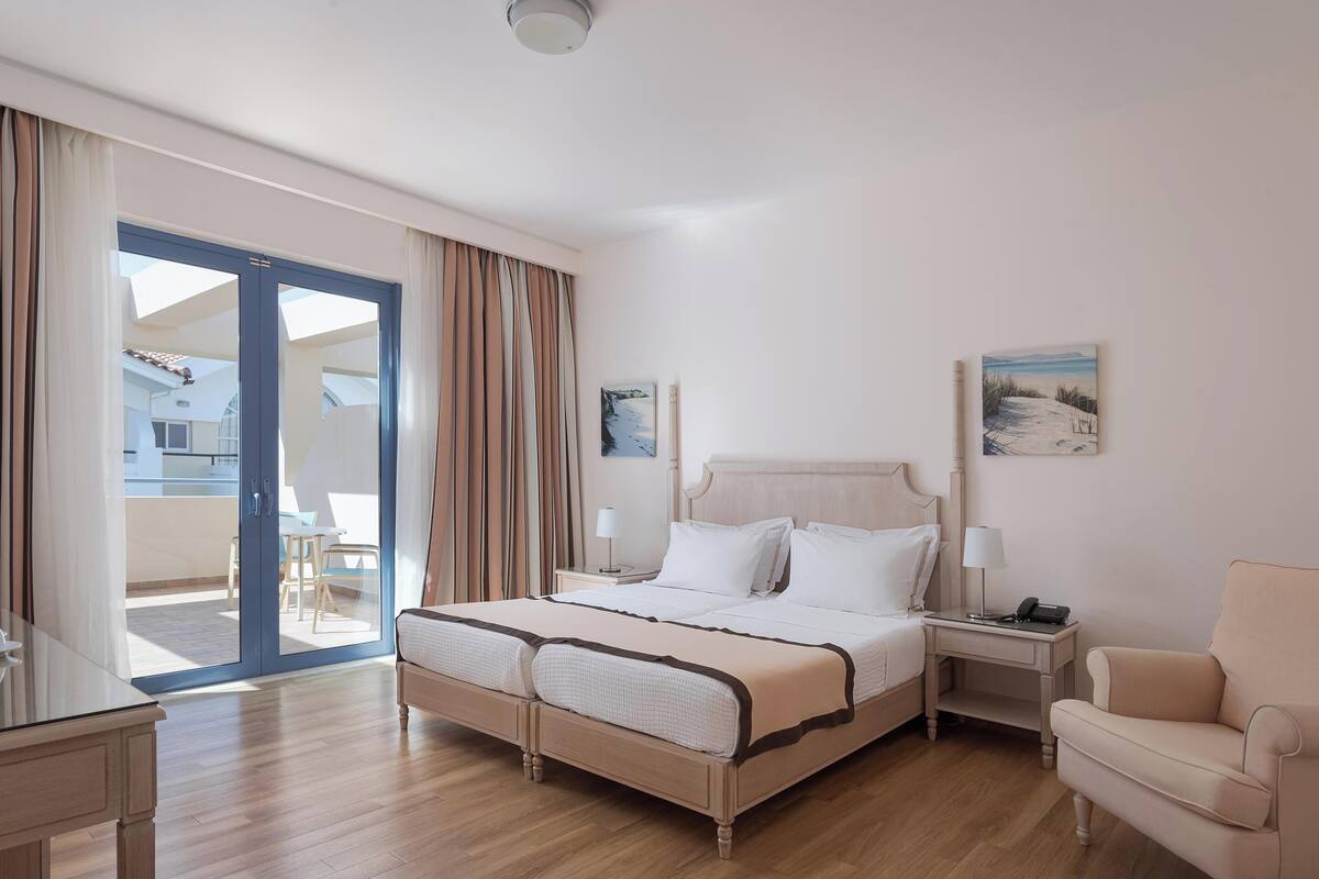 A hotel room with a large bed, a desk, a chair, and a balcony with curtains.