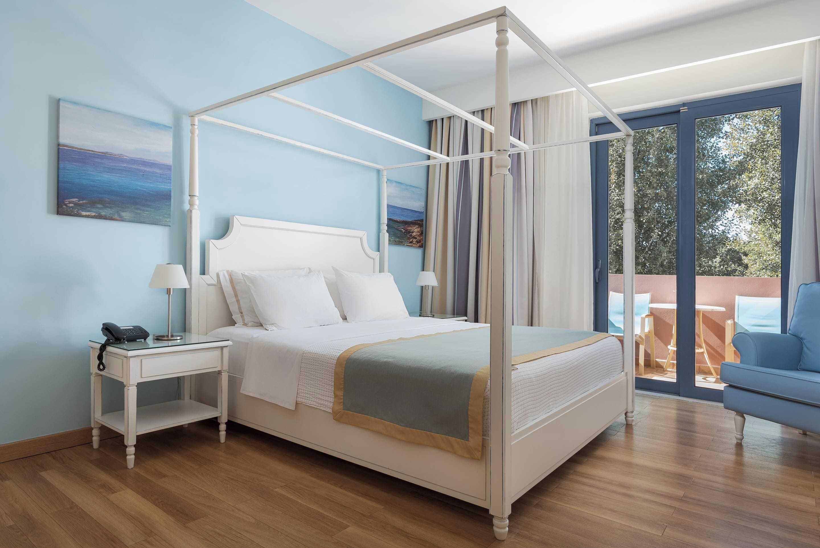 A four-poster bed with a white frame, a bedside table with a lamp, and a view of the outdoors through a large window.
