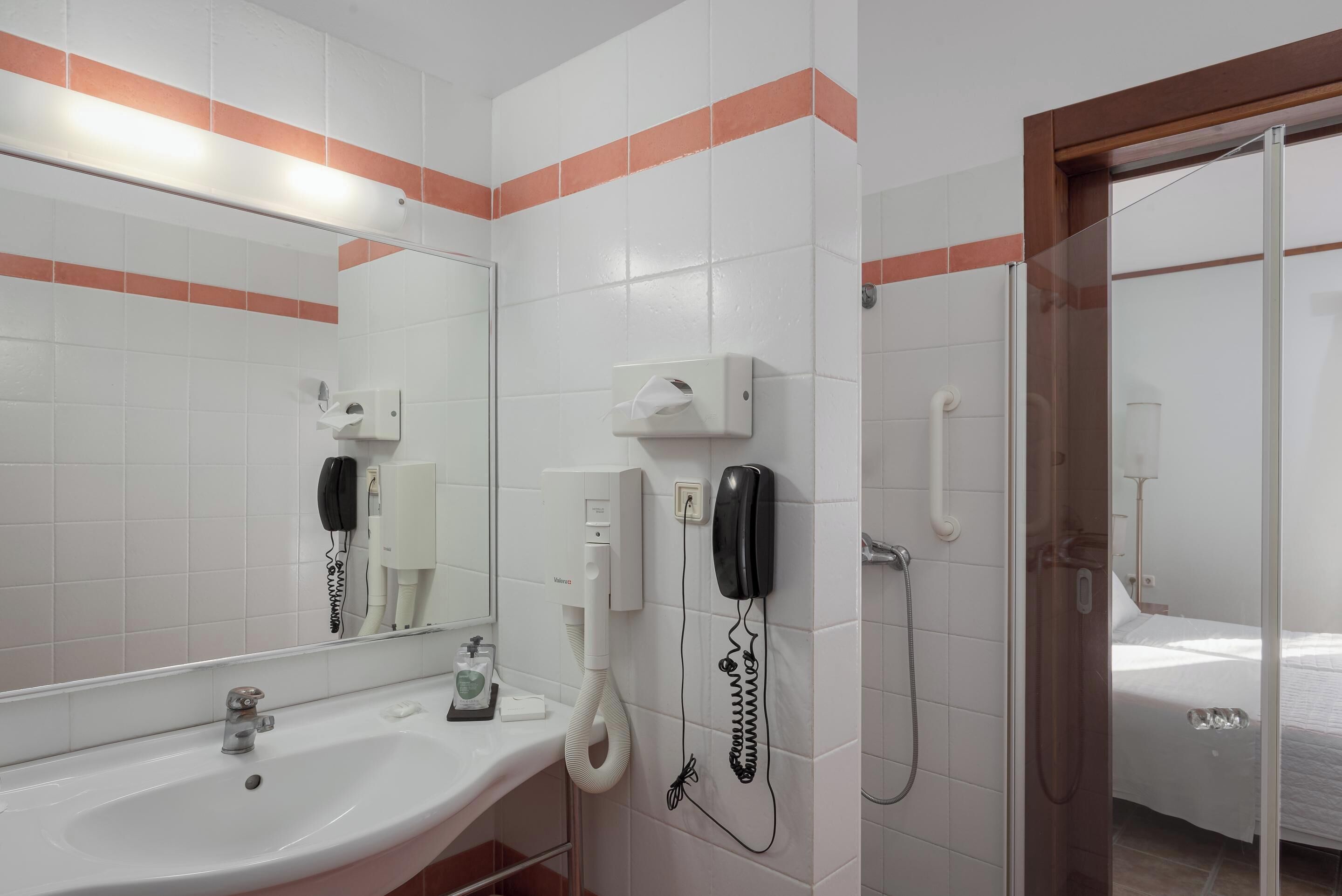 Casa de banho de hotel com lavatório, espelho, dispensador de toalhas, telefone e chuveiro.