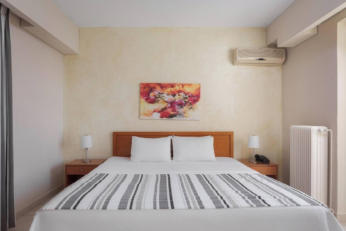 A hotel room with a bed, two bedside tables, a wall-mounted air conditioner, a radiator, and a painting on the wall.