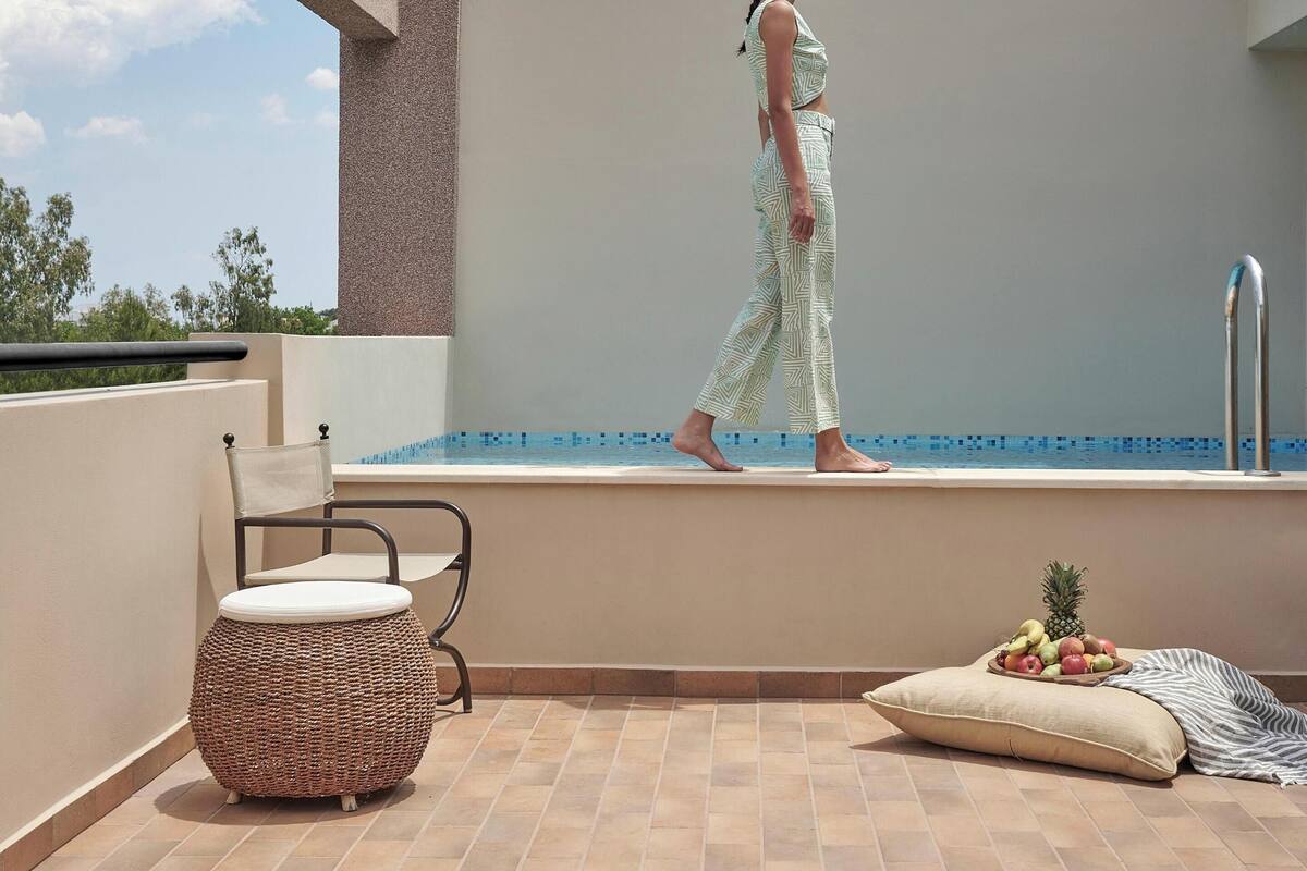 A person walking on a balcony with a pool, a wicker chair, and a fruit basket.