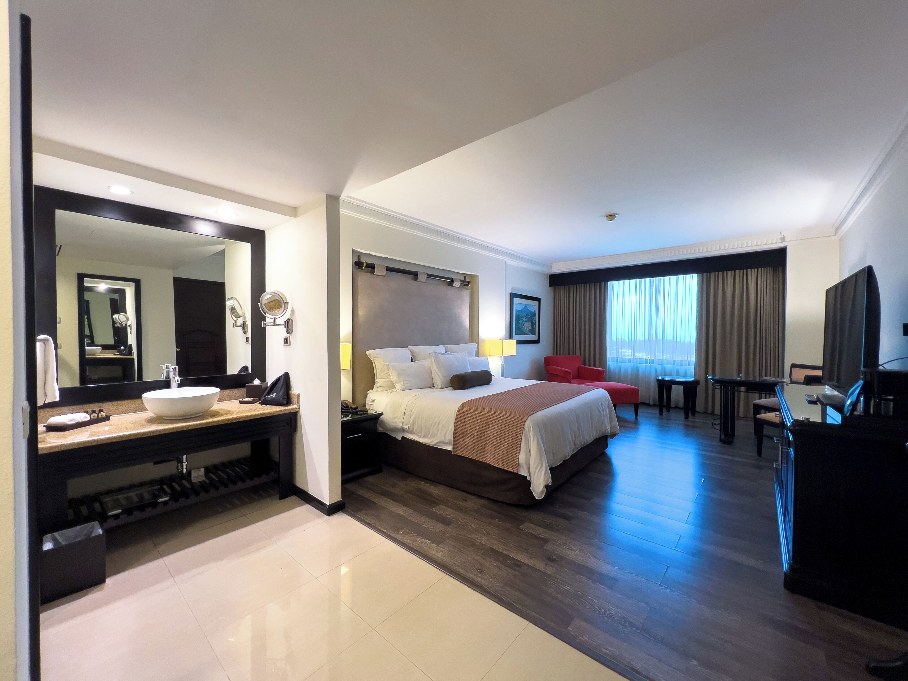A hotel room with a large bed, a desk, a TV, a bathroom with a sink and mirror, and a view of the outdoors.