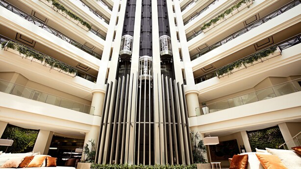 A multi-story hotel atrium with a central column, balconies with plants, and seating areas with cushions.