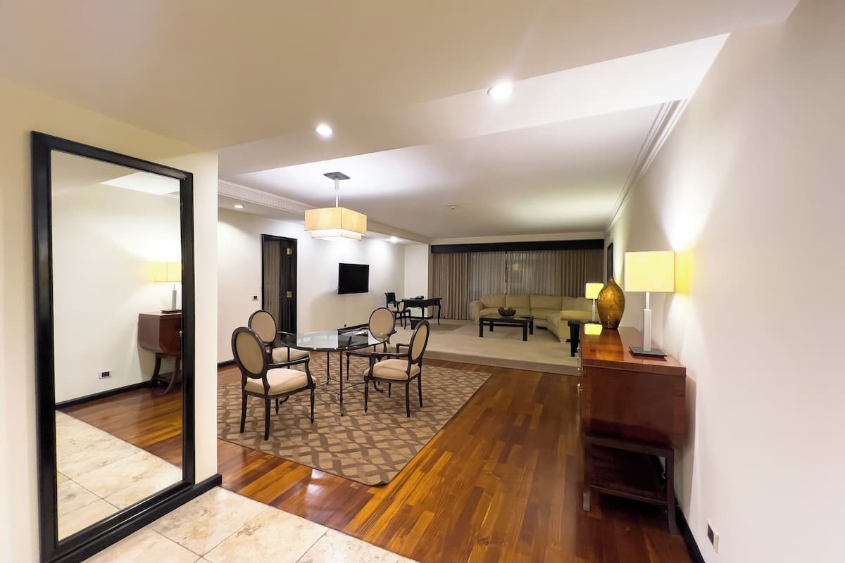 Amplia habitación de hotel con un espejo grande, un área de comedor con una mesa y sillas de vidrio, un sofá y un televisor montado en la pared.