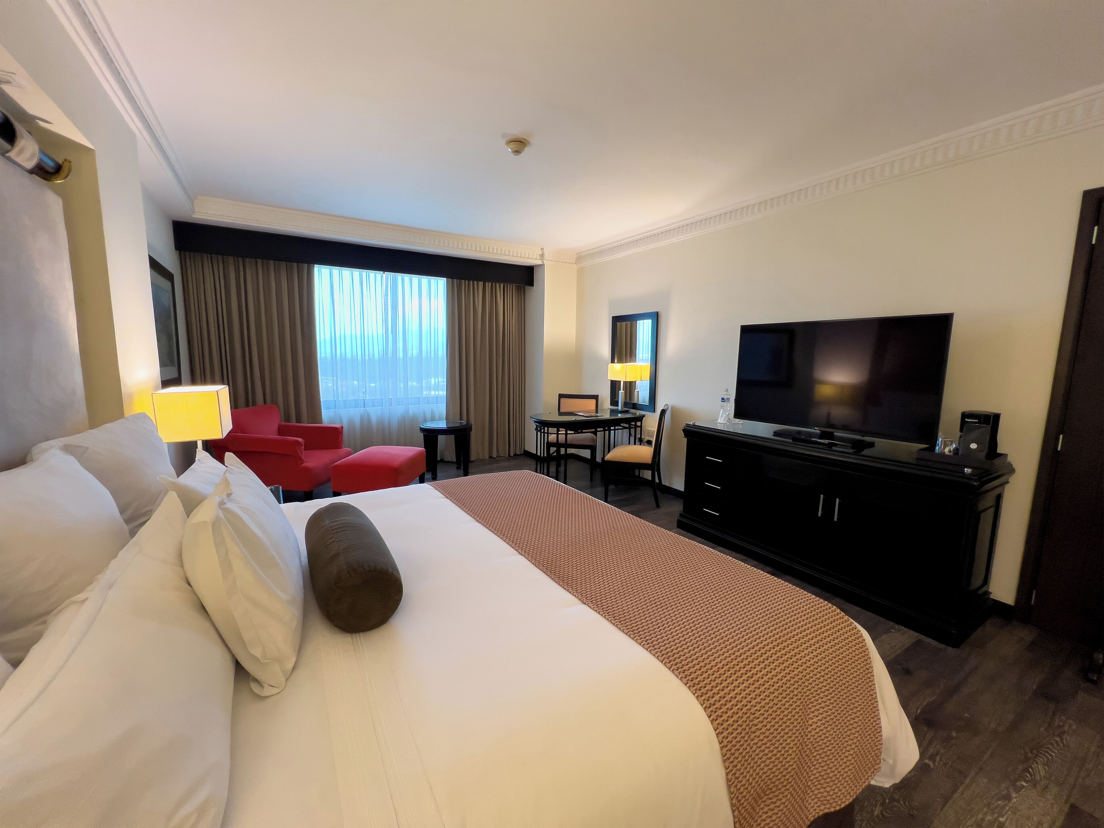 Une chambre d’hôtel avec un grand lit, un bureau avec un ordinateur, une télévision et un fauteuil rouge.