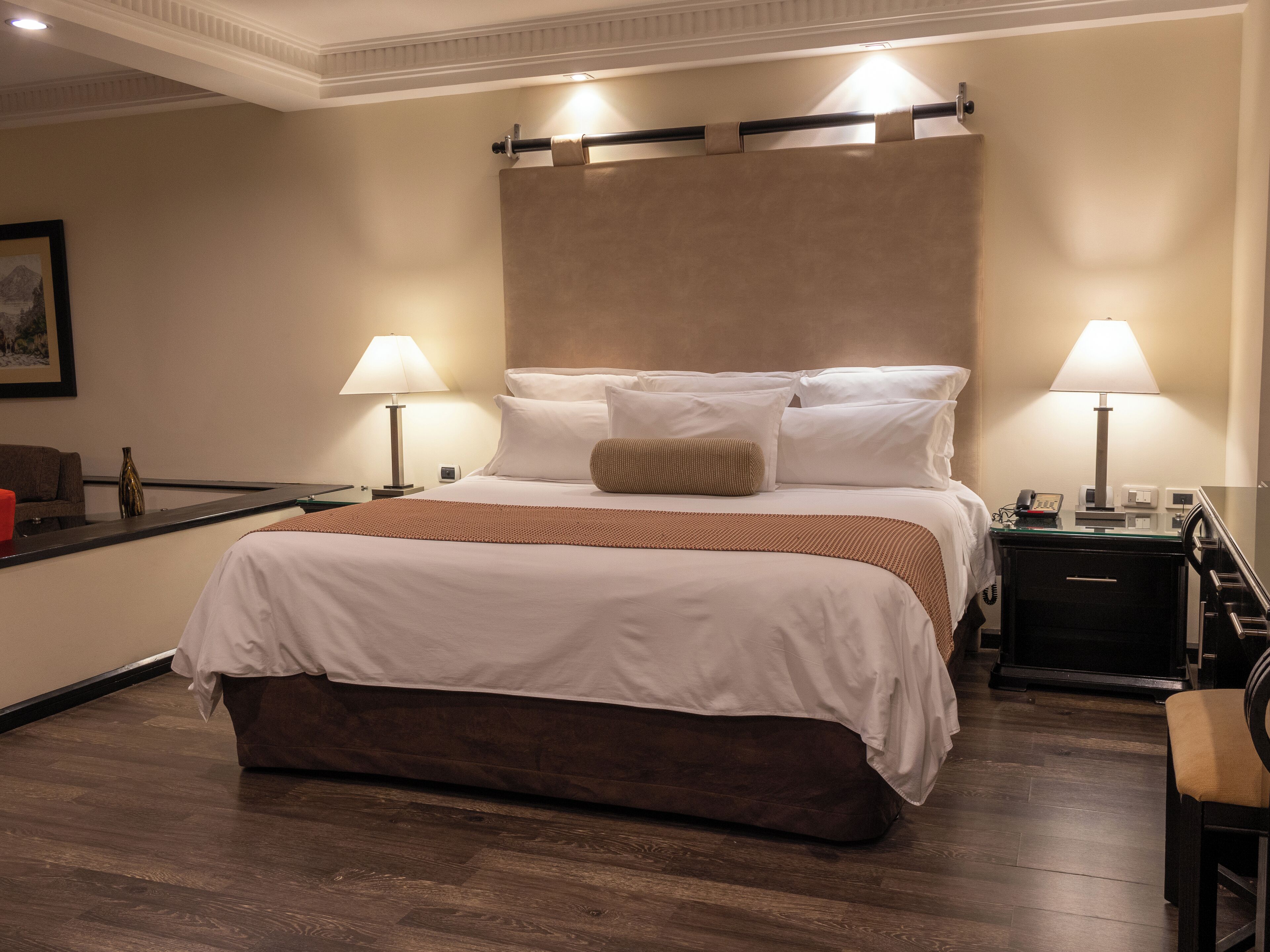 Quarto de hotel com cama grande, duas luminárias de cabeceira, mesa de cabeceira, escrivaninha e uma cadeira.