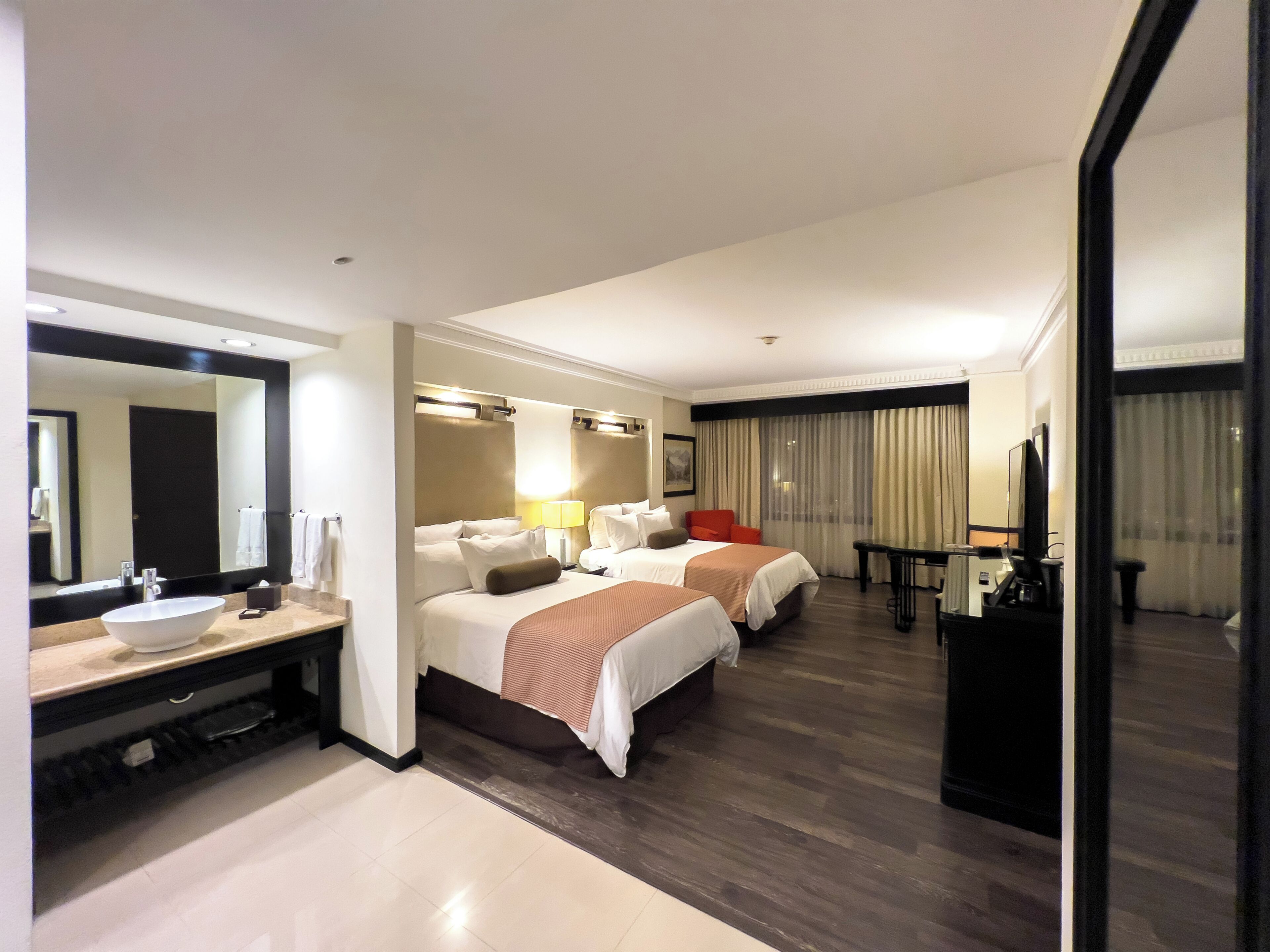 A hotel room with a large bed, a smaller bed, a desk, a chair, a TV, a mirror, and a bathroom with a sink and mirror.