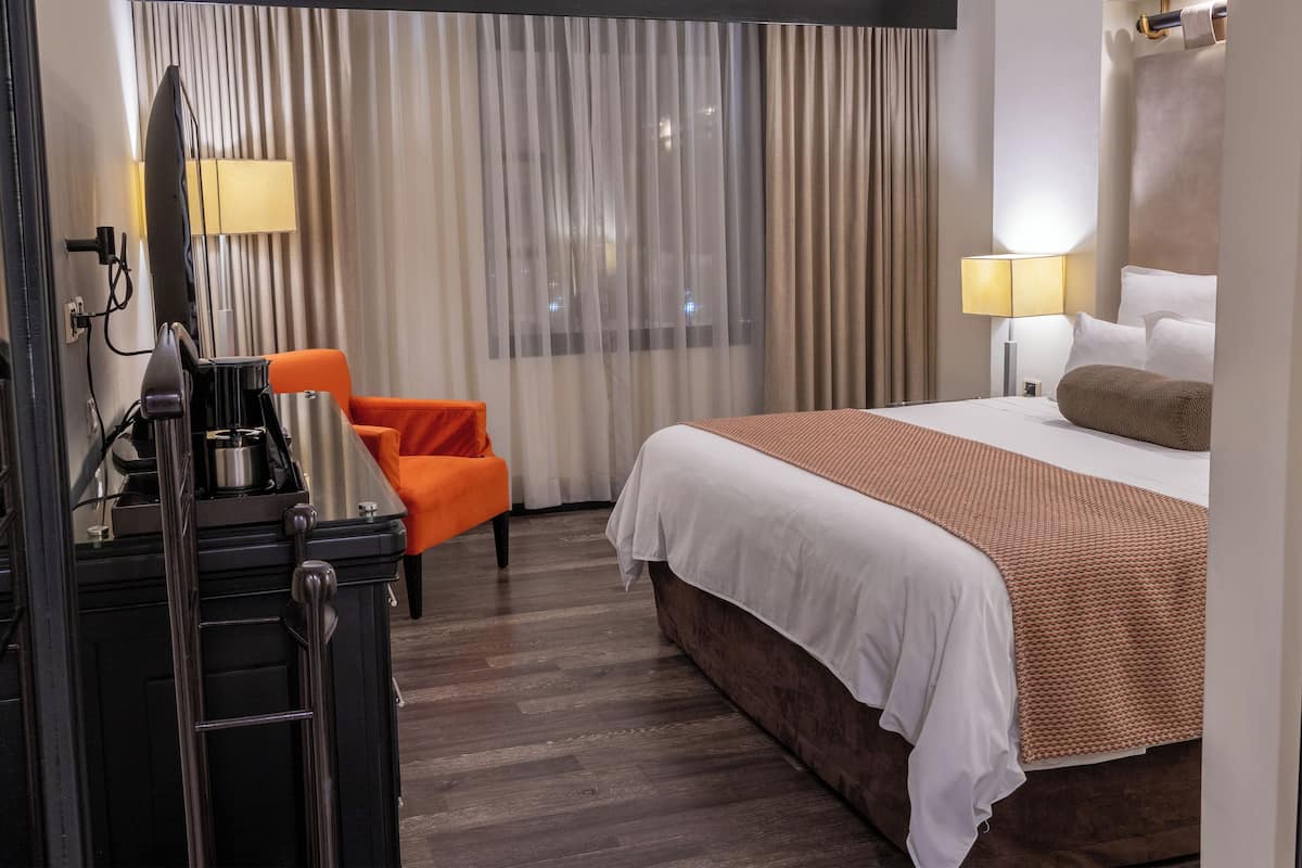 Habitación de hotel con cama, escritorio con cafetera, una silla naranja, ventana con cortinas y una lámpara.