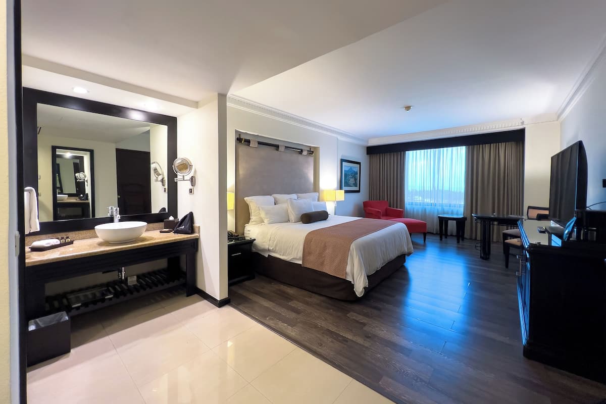 Habitación de hotel con una cama grande, zona de estar con televisor, escritorio y baño con lavabo y espejo.
