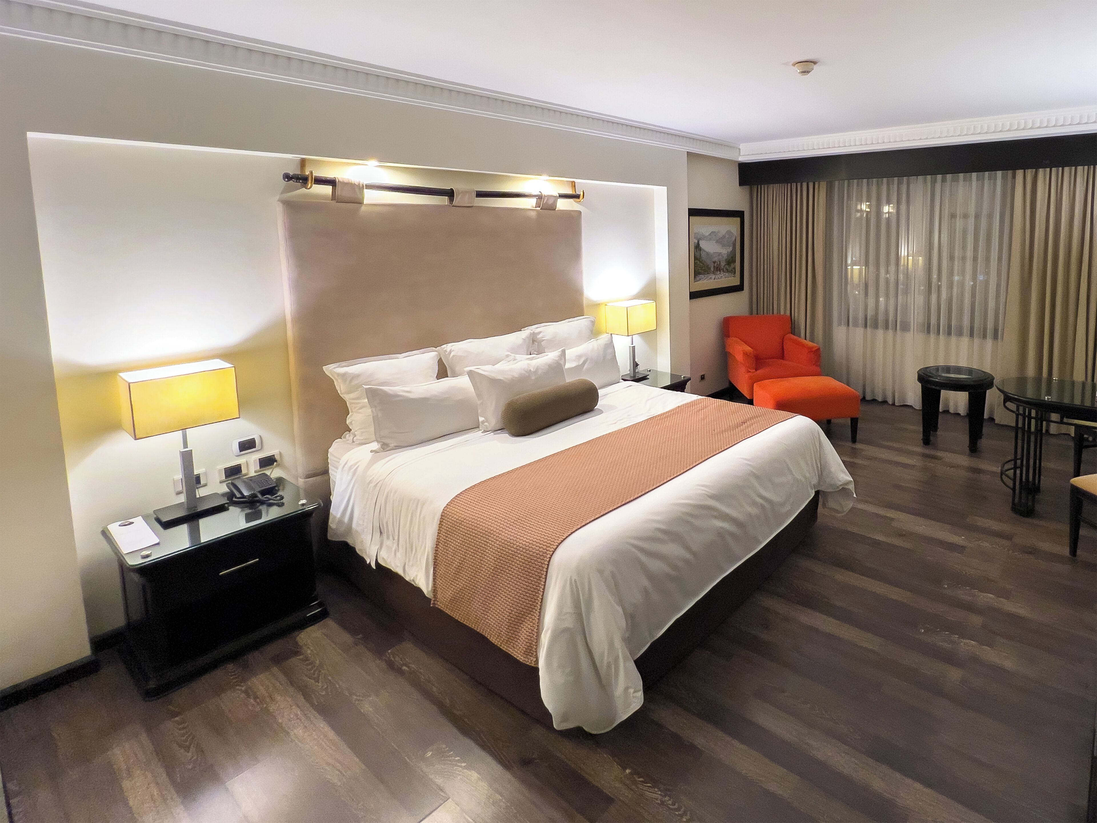 A hotel room with a large bed, a nightstand with a lamp, a chair, a small table, and a window with curtains.