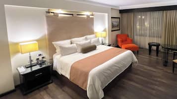 A hotel room with a large bed, a nightstand with a lamp, a chair, a small table, and a window with curtains.
