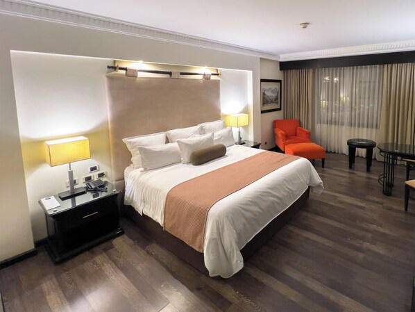 Una habitación de hotel con una cama grande, una mesita de noche con lámpara, una silla, una pequeña mesa y una ventana con cortinas.