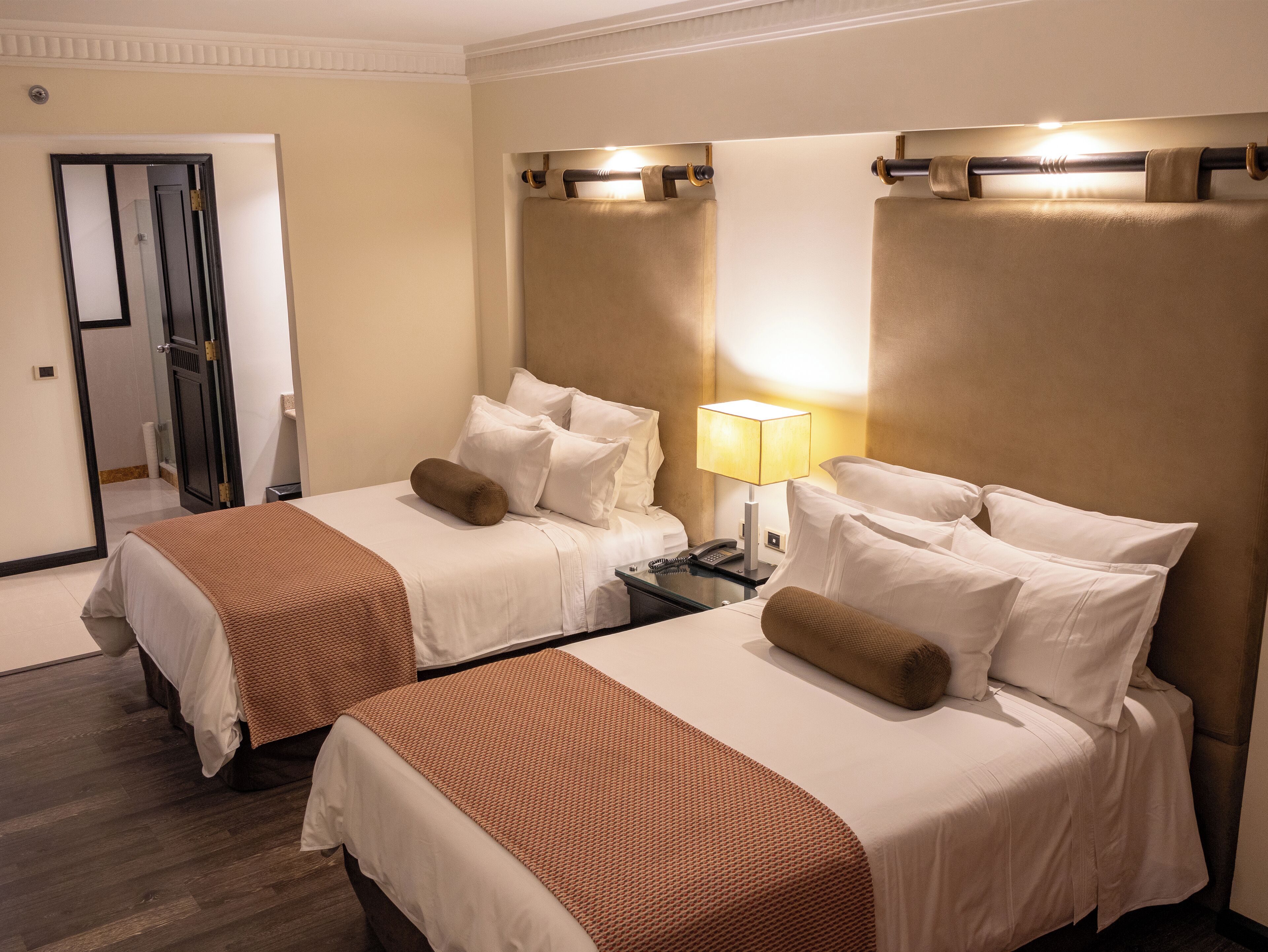 Habitación de hotel con dos camas, cada una con sábanas blancas y almohadillas marrones, una mesita de noche con lámpara y un espejo en la pared.