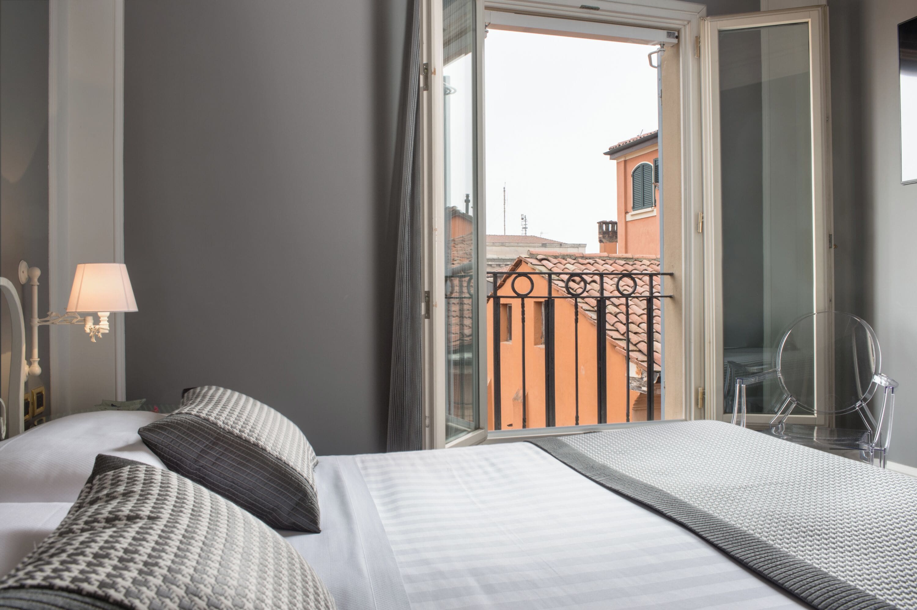 Habitación de hotel con una cama, una mesita de noche, una lámpara y vistas a un balcón con barandilla.