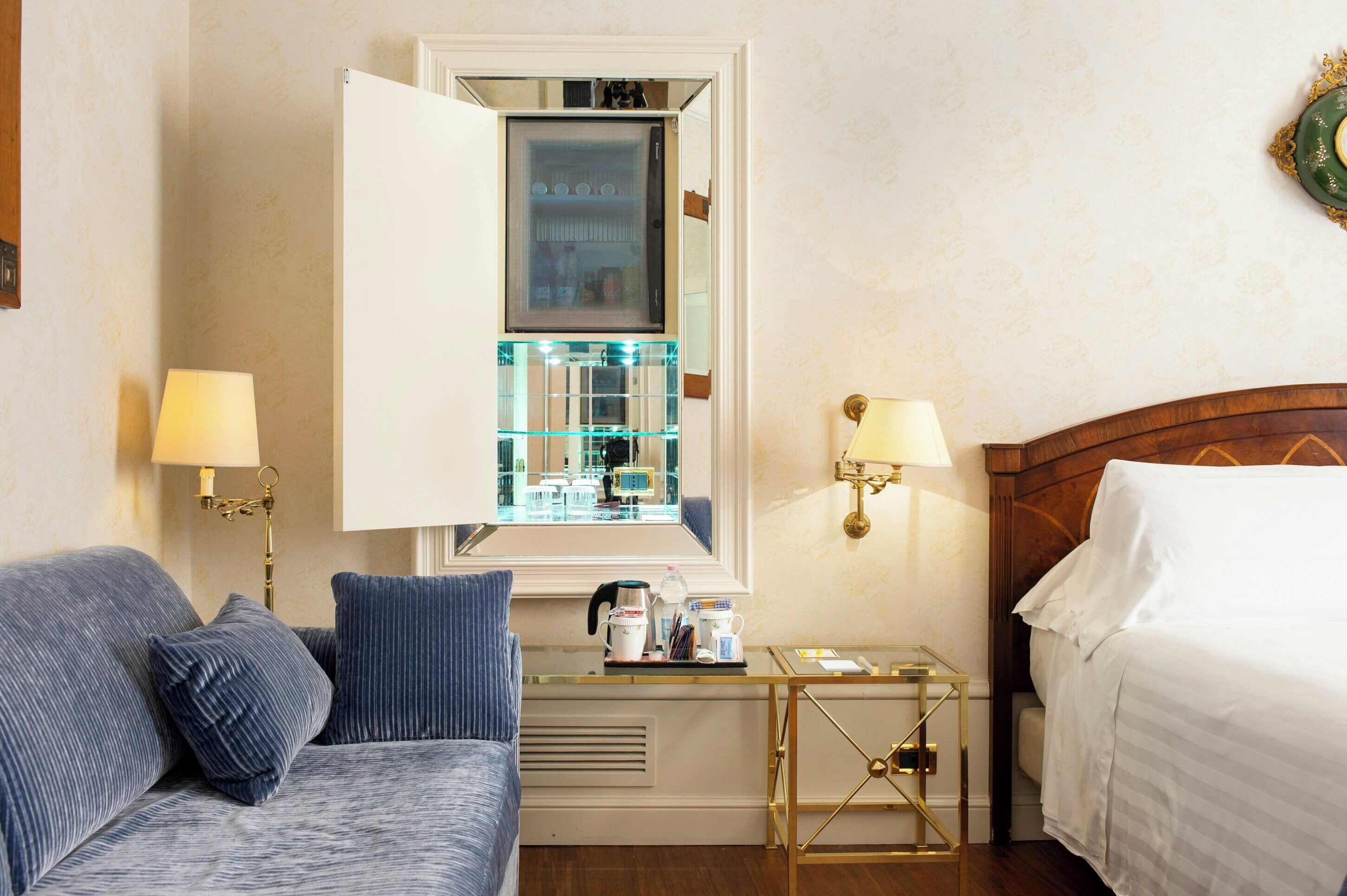 A hotel room with a bed, a sofa, a small table with a lamp, and a mirror reflecting the room.