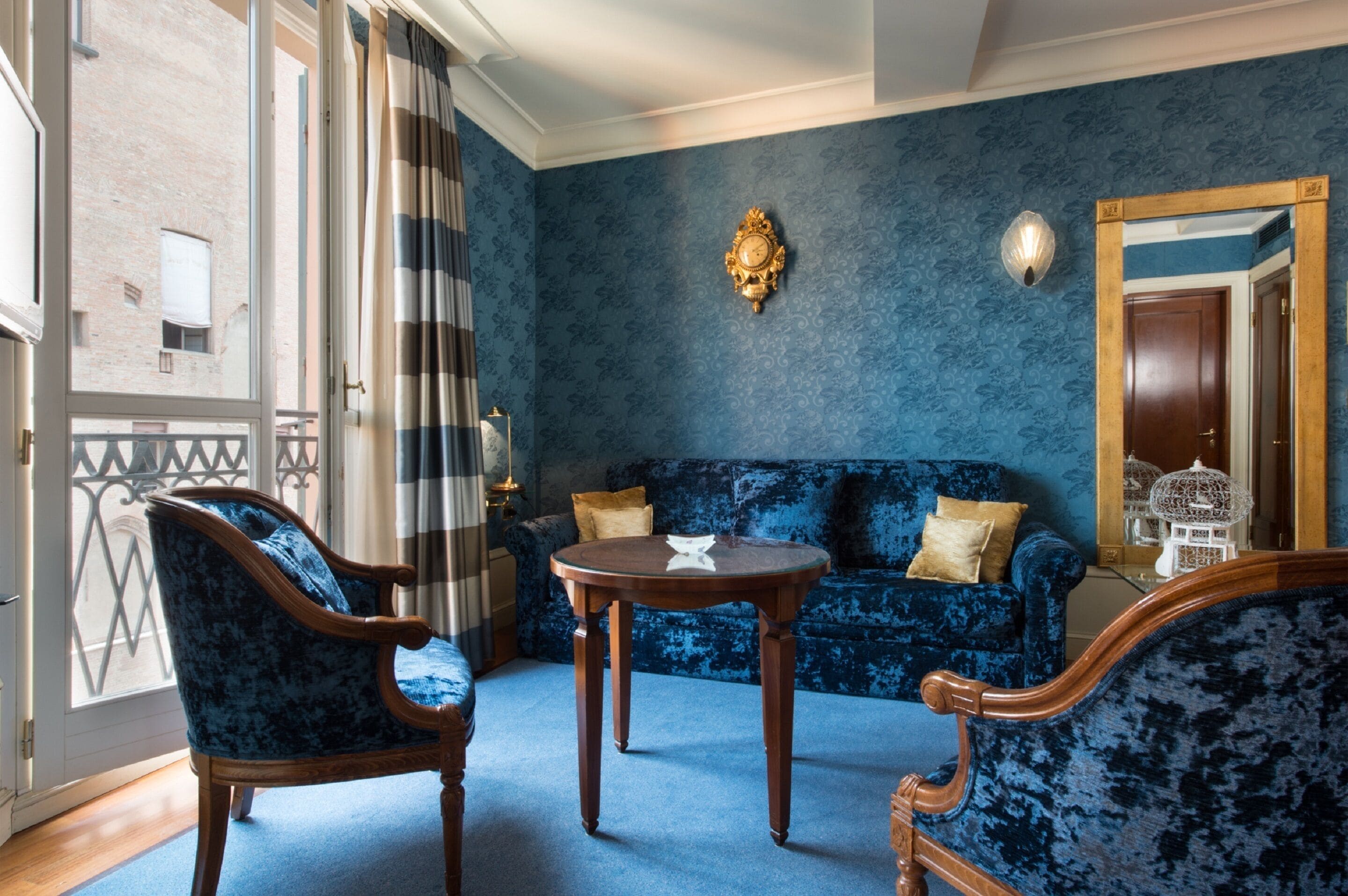 A room with a blue patterned wall, a wooden sofa, a round wooden table, and a large mirror.